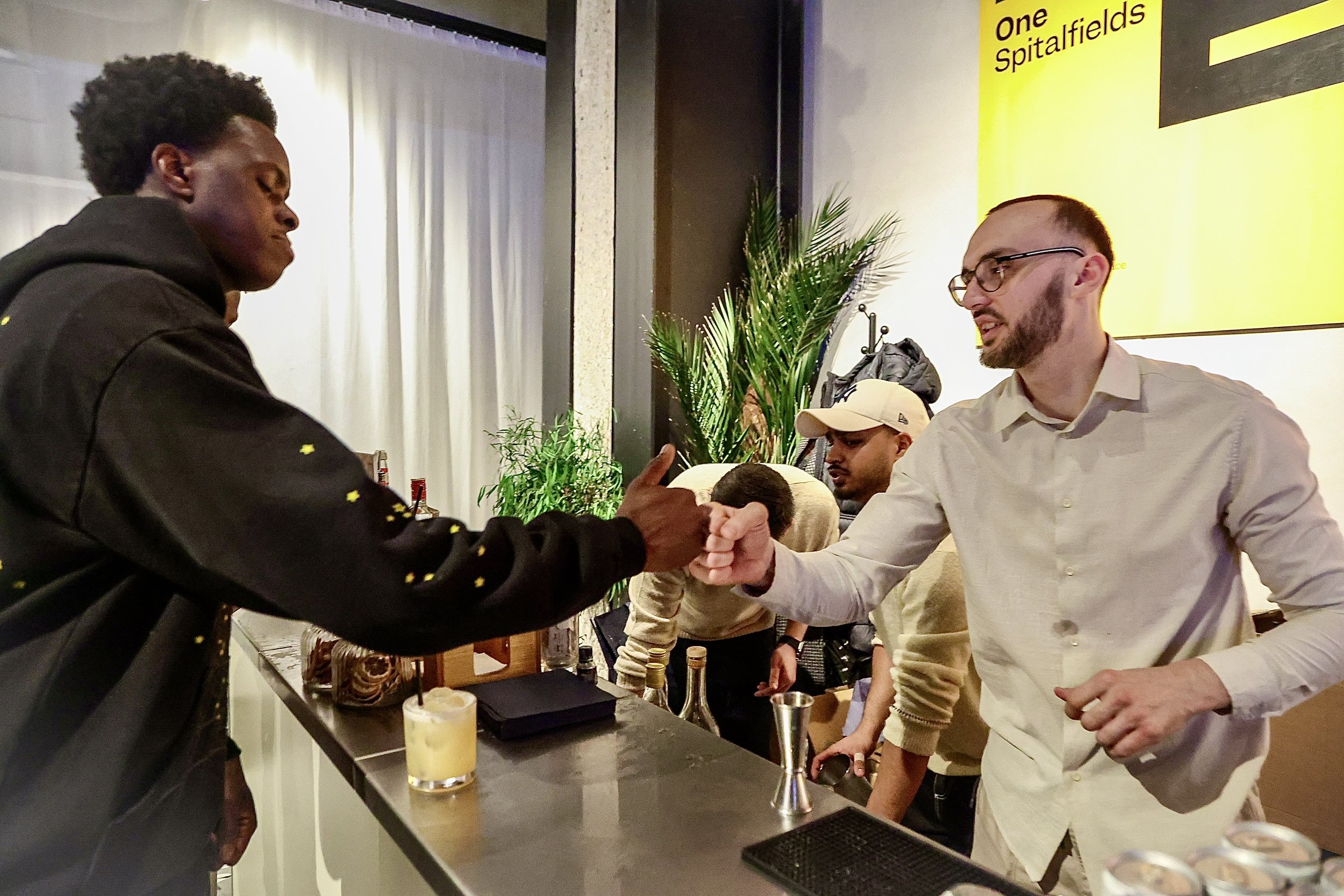 Two men at a bar giving a fist bump, one wearing a black hoodie, and the other wearing a beige shirt and glasses. There are plants and other people in the background.