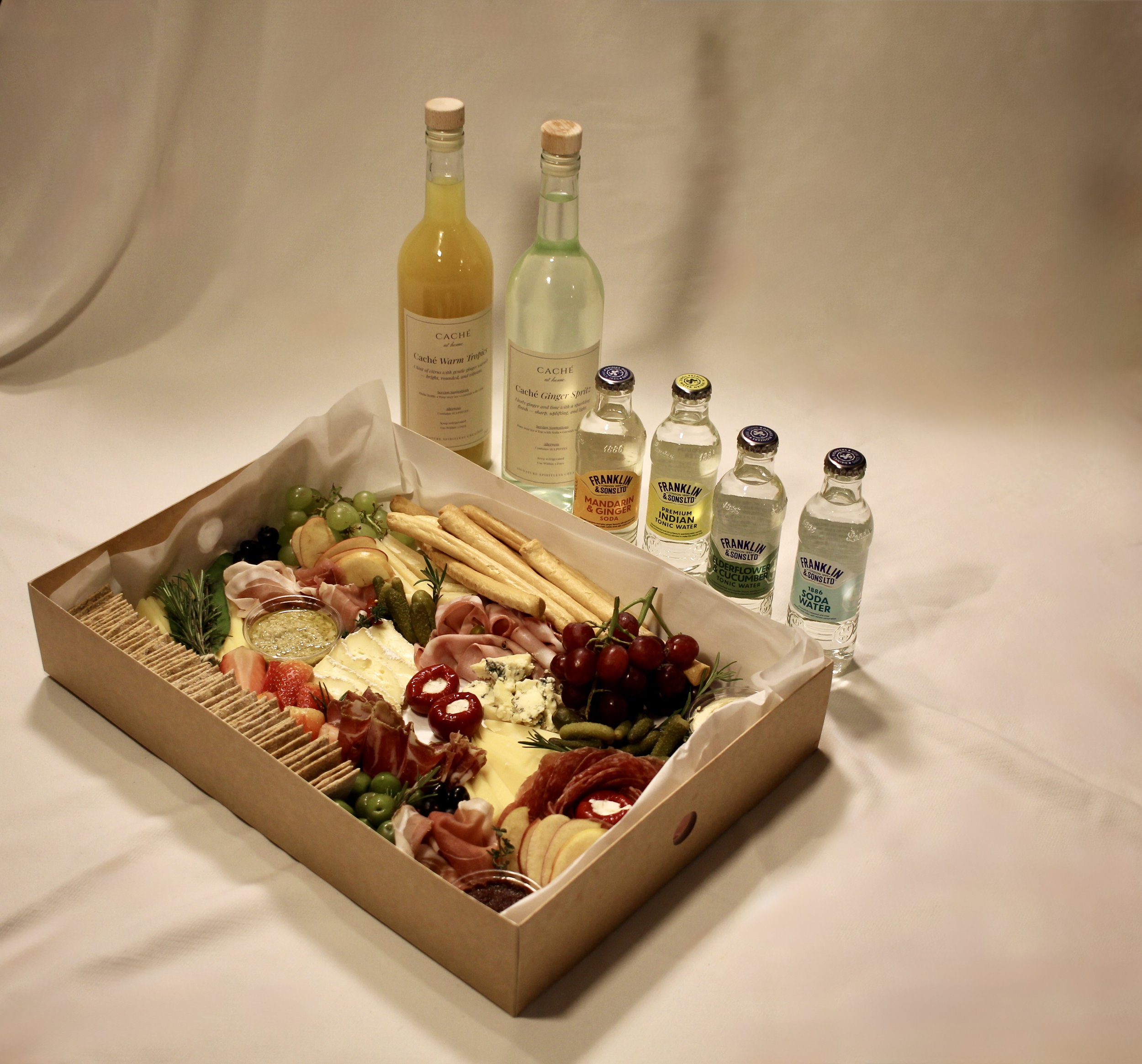 A charcuterie platter with assorted meats, cheeses, fruits, and crackers, accompanied by bottles of sparkling water and flavored syrups on a white table.