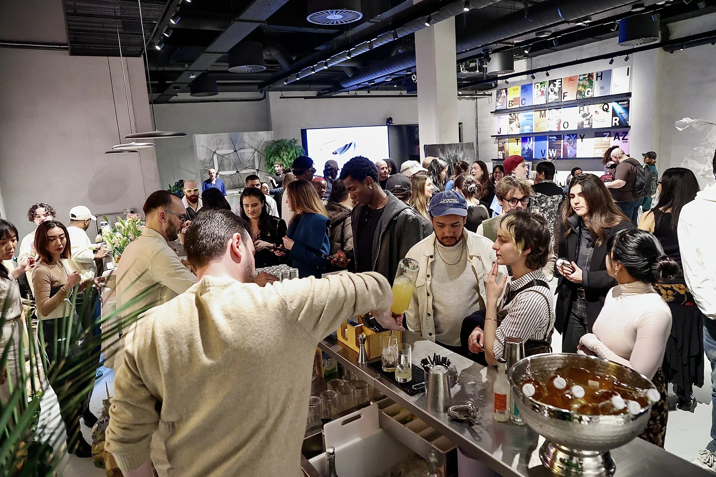 A crowded indoor event with people gathered around a bar where a bartender is pouring a yellowish drink. Many people are talking, some holding drinks, in a modern space with colorful artwork and alphabet-themed decor on the walls.