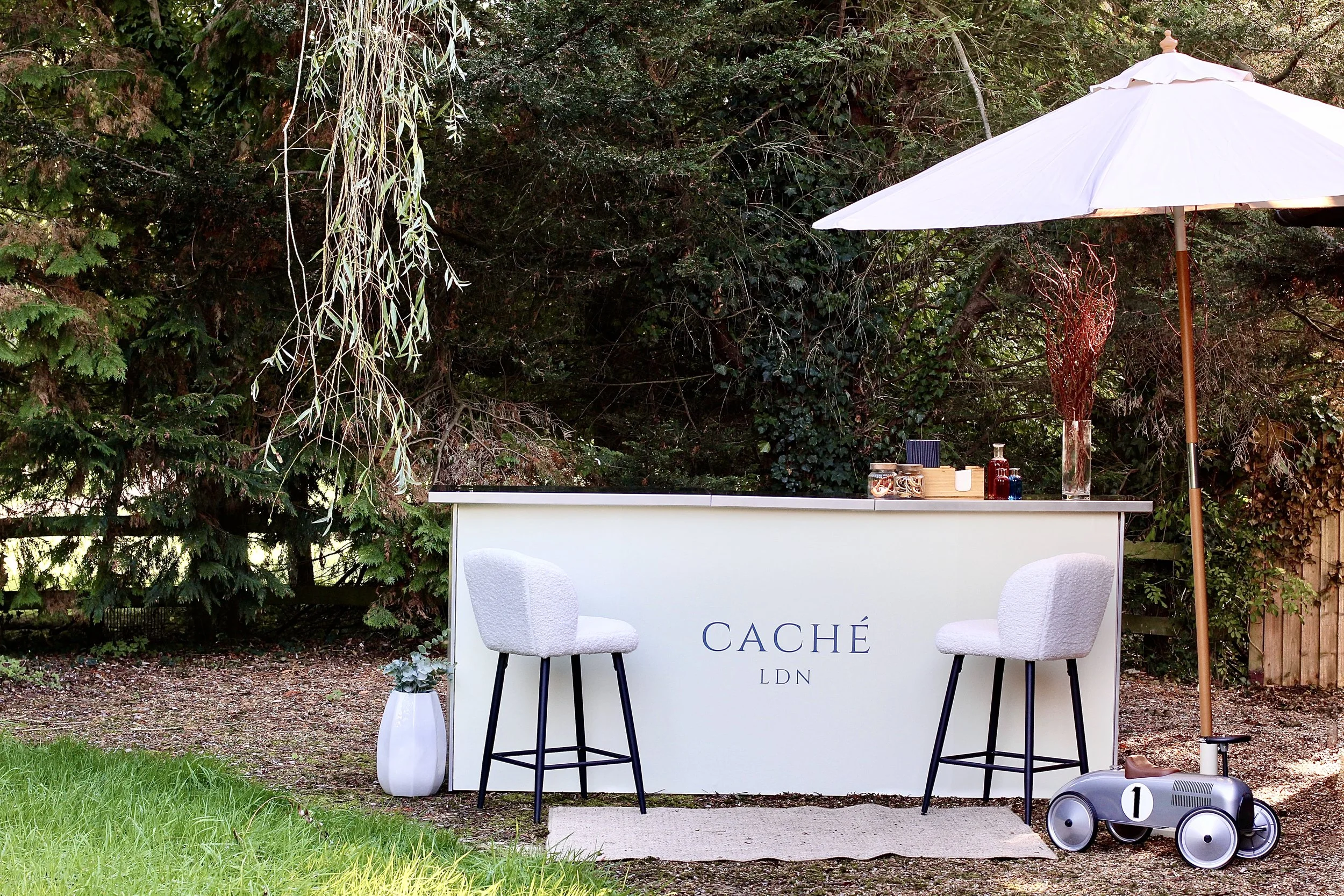 Outdoor bar setup with a white counter labeled 'CACHÉ LDN', two white bar stools, a large white patio umbrella, a decorative plant in a white vase, and a toy ride-on car for children, with trees and greenery in the background.