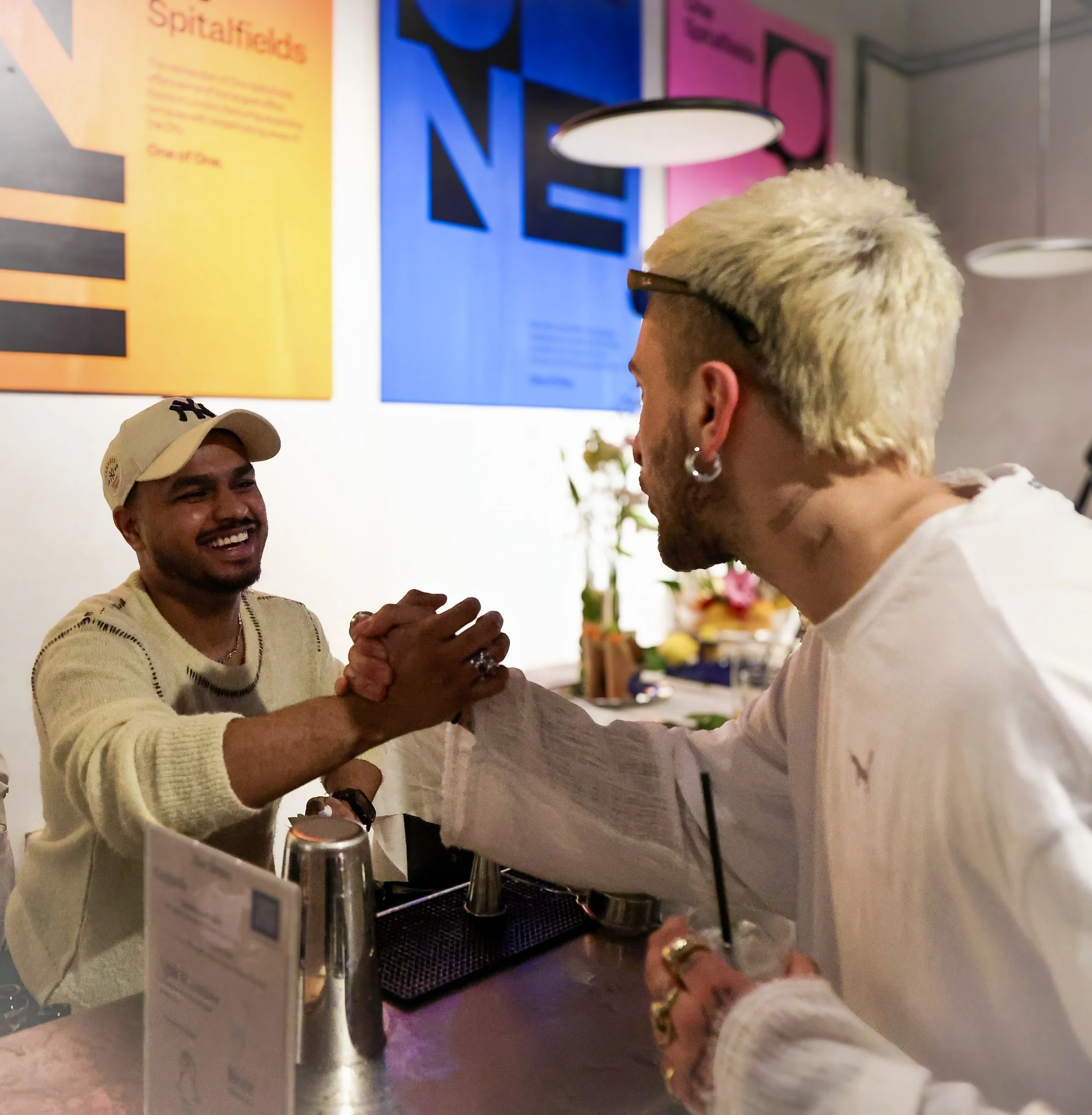 Two men shaking hands in a bar or restaurant, smiling at each other, with posters on the wall behind them.