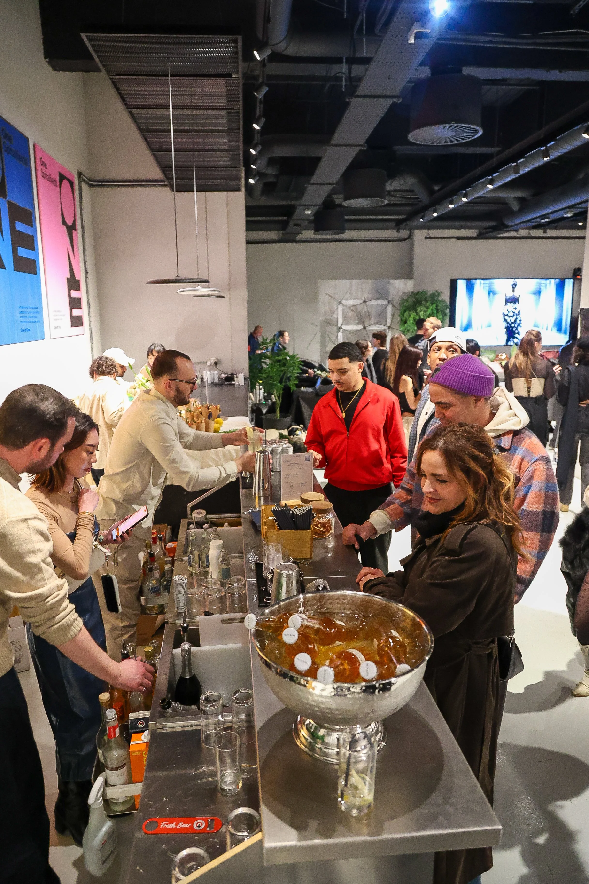 People gathered around a bar at an event or party, with bottles and glasses on the counter, and a large screen in the background showing a fashion show.