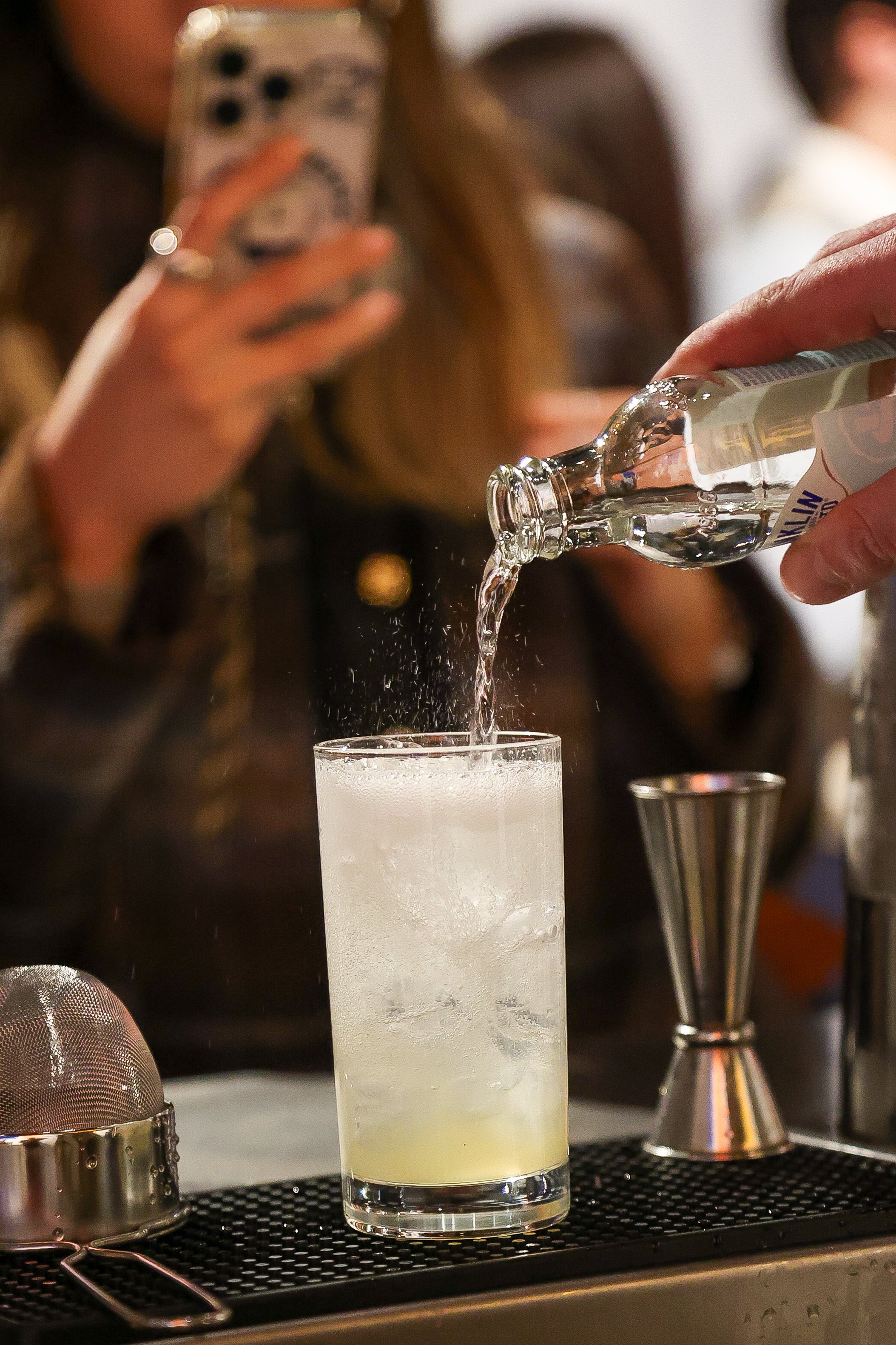 A bartender pours clear liquor into a tall glass with ice, while a woman in the background takes a photo with her phone.