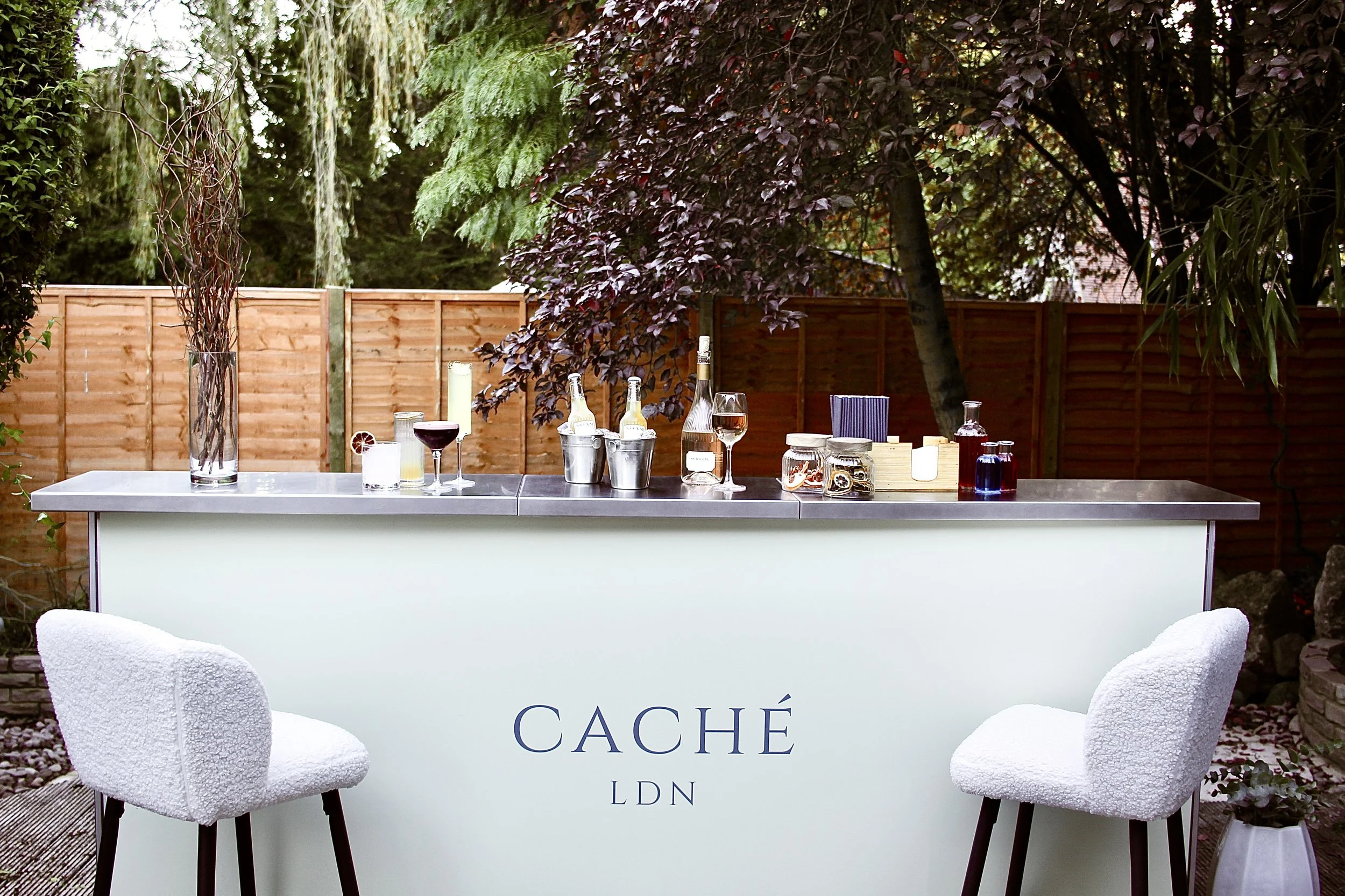 Outdoor bar with two white fuzzy chairs, various bottles and glasses, and a wooden fence in the background.