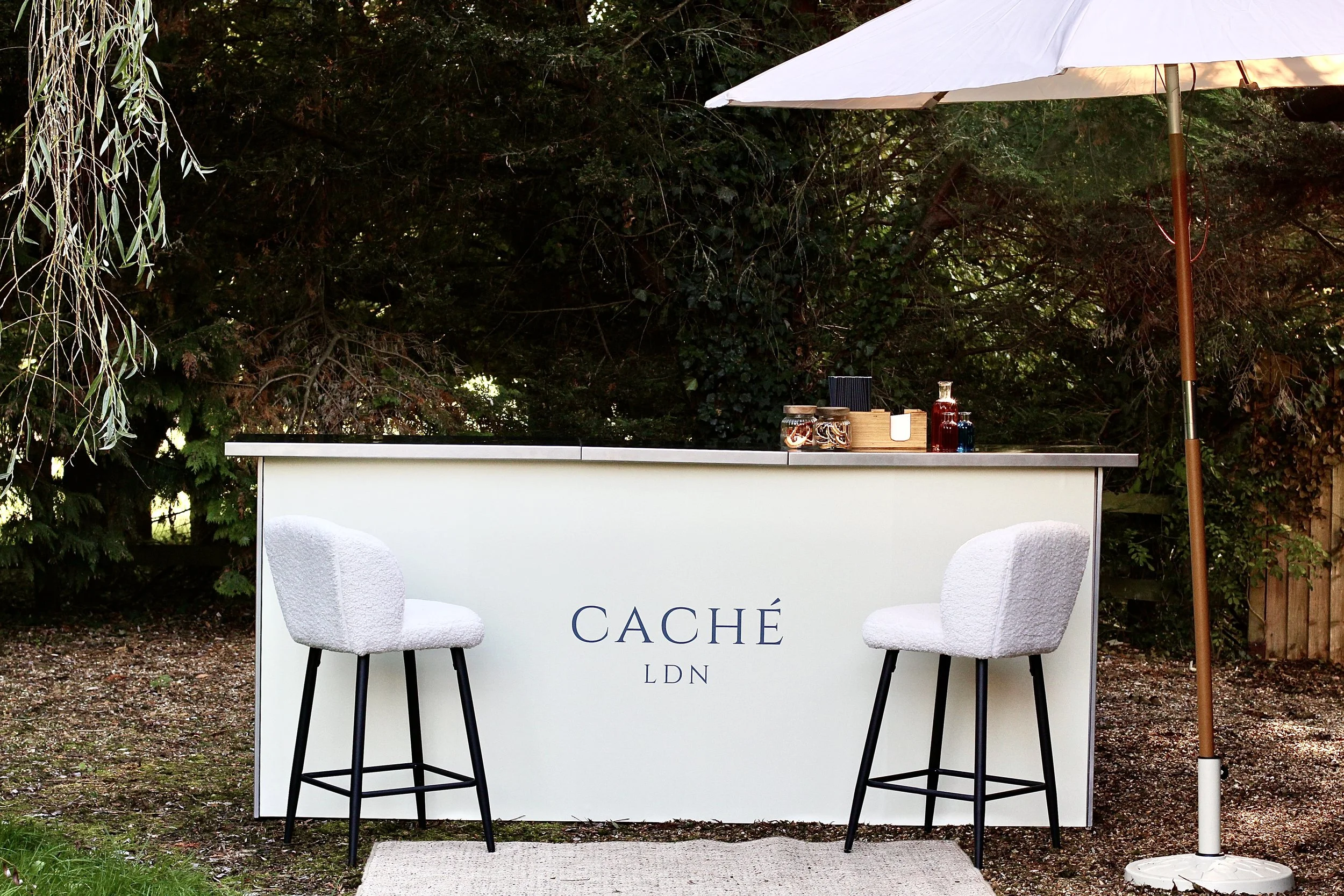 Outdoor bar with two white chairs, a white counter with the word 'CACHÉ LDN' printed on it, and an umbrella on the right. The bar is set against a backdrop of greenery.