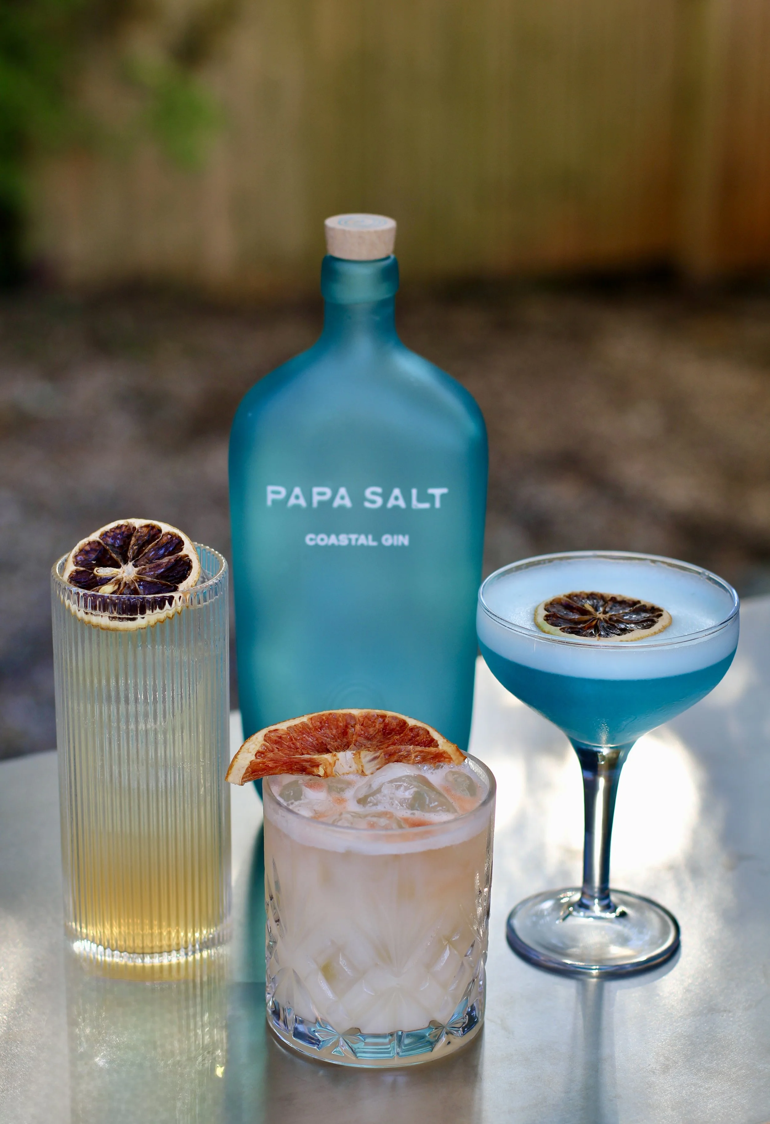 Three colorful cocktails with dried citrus garnishes in front of a blue bottle labeled 'PAPA SALT COASTAL GIN' on a white surface outdoors.