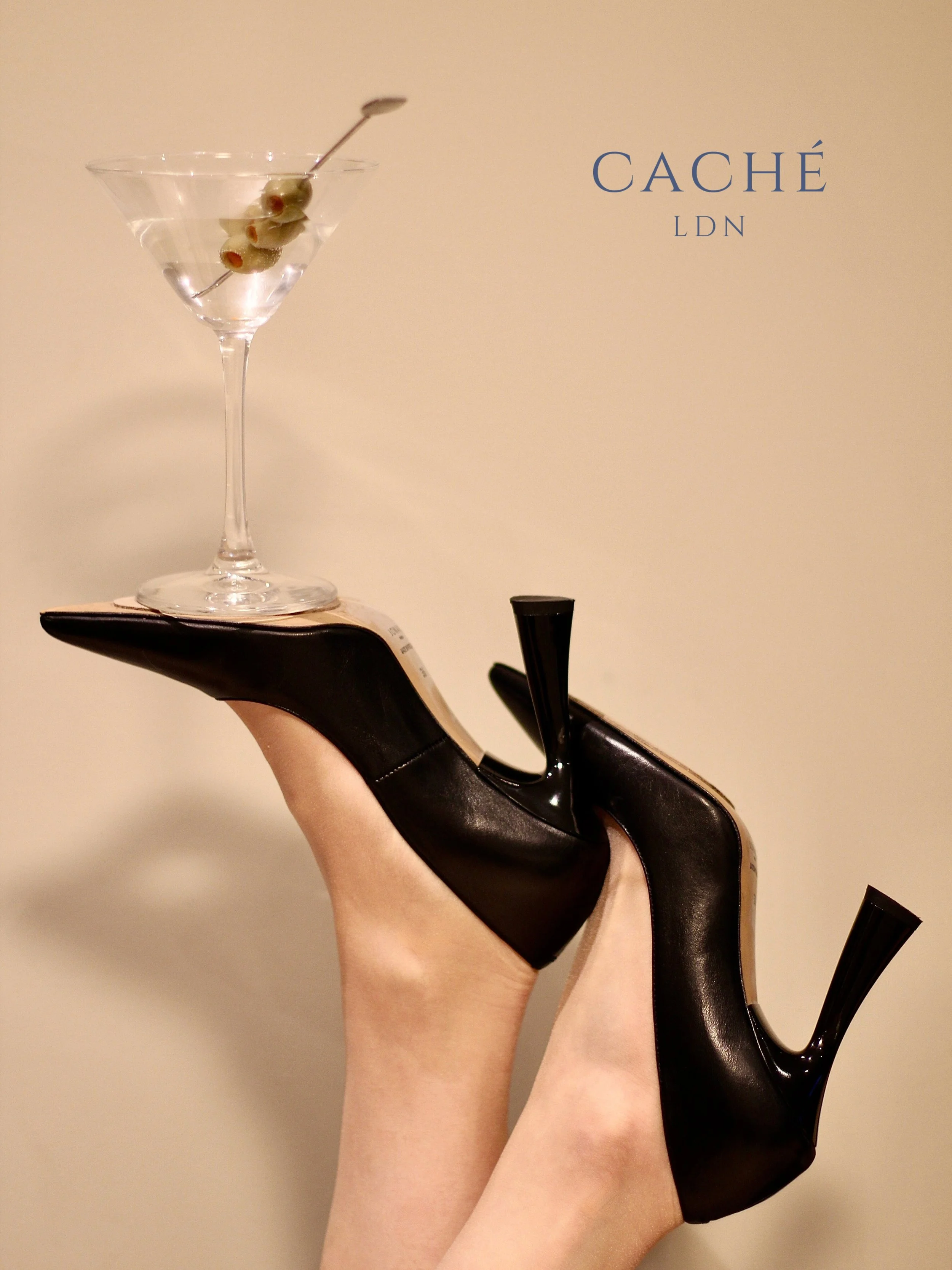 A person wearing black high heels with their feet up, holding a martini glass with green olives on a skewer. The background features the text 'CACHÉ LDN'.