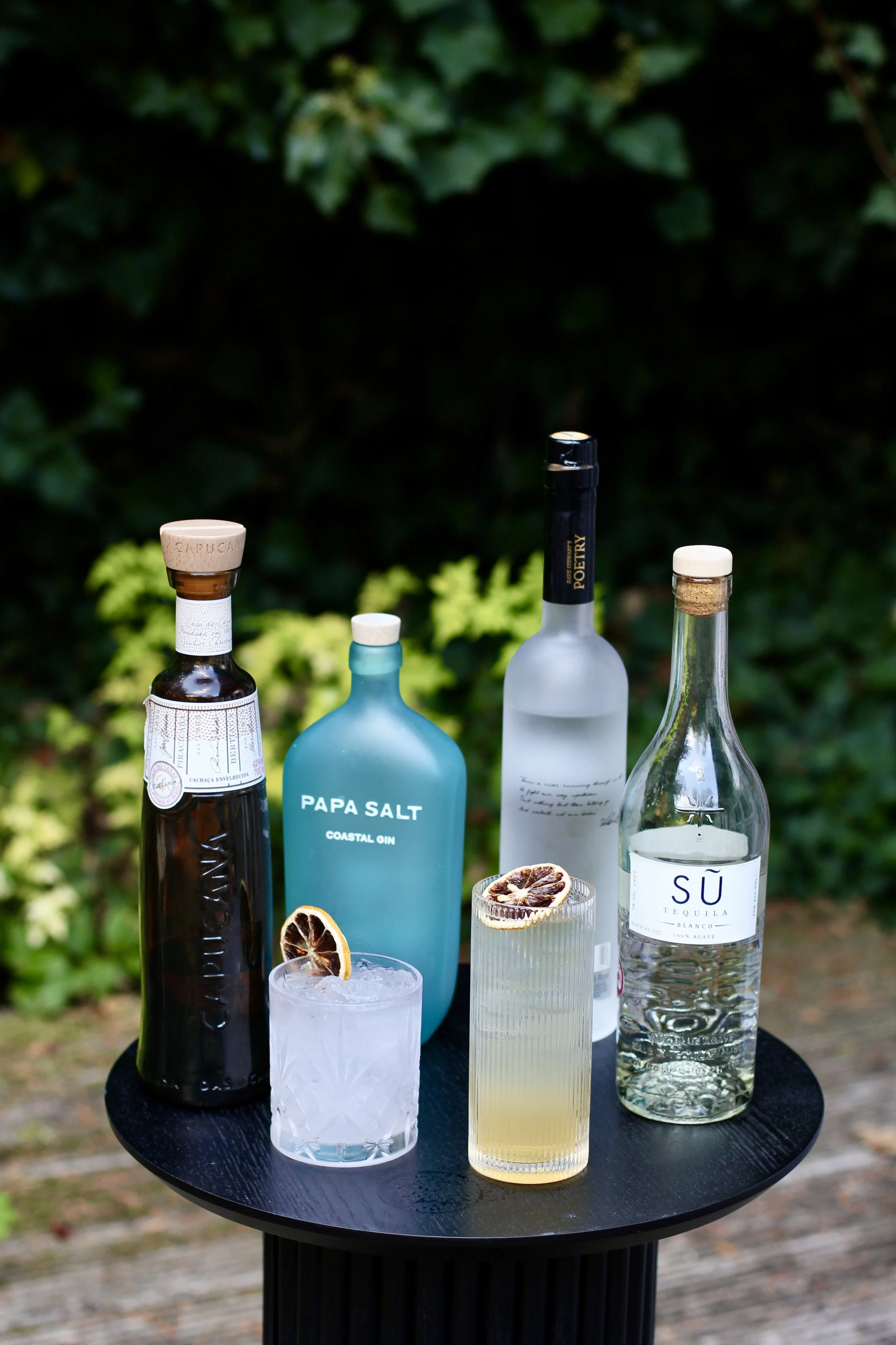 Assorted bottles of alcohol and two cocktails with dried citrus slices on a black round table outdoors.