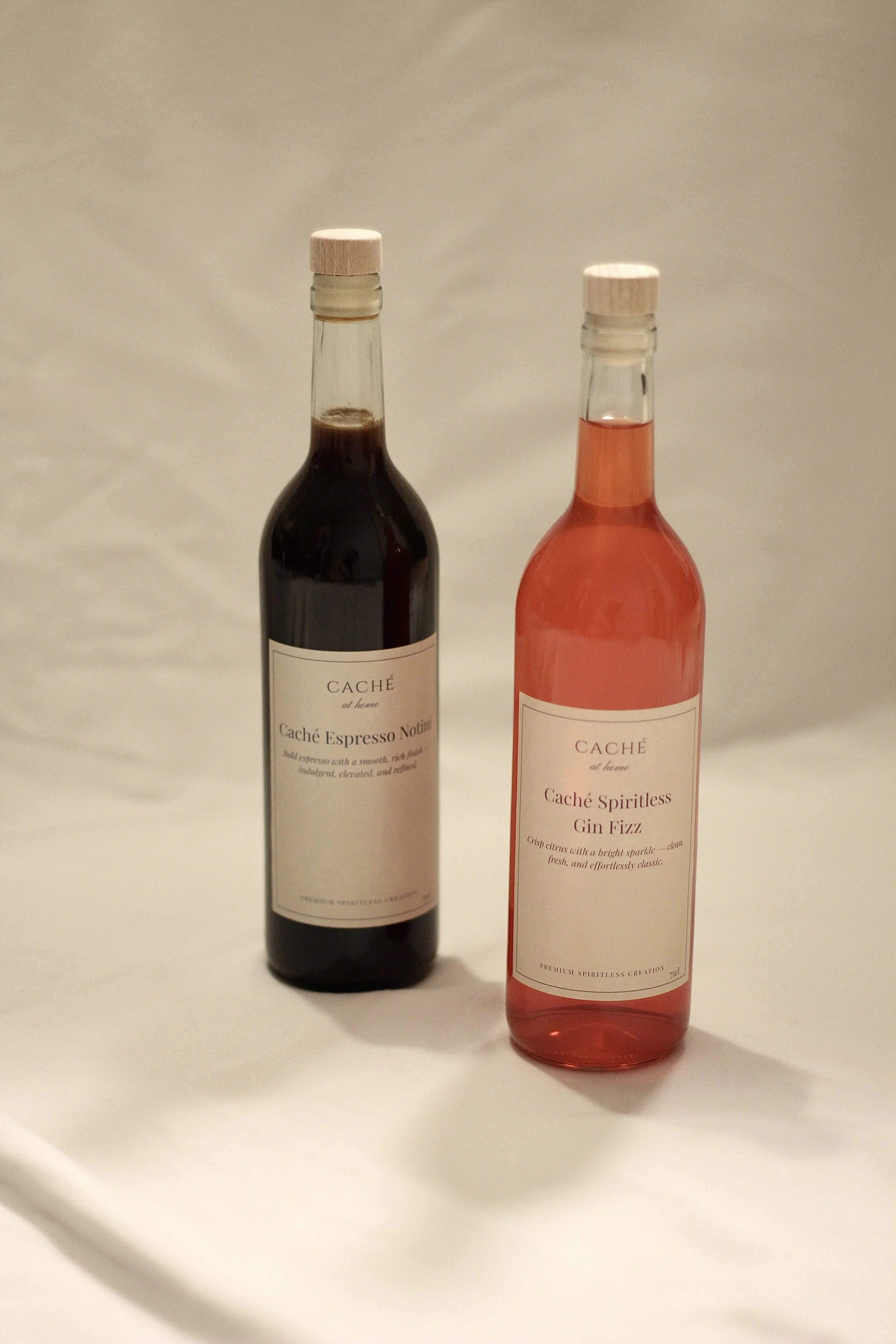 Two bottles of flavored spirits on a white surface, one dark and one pink, with labels indicating espresso and gin fizz flavors.