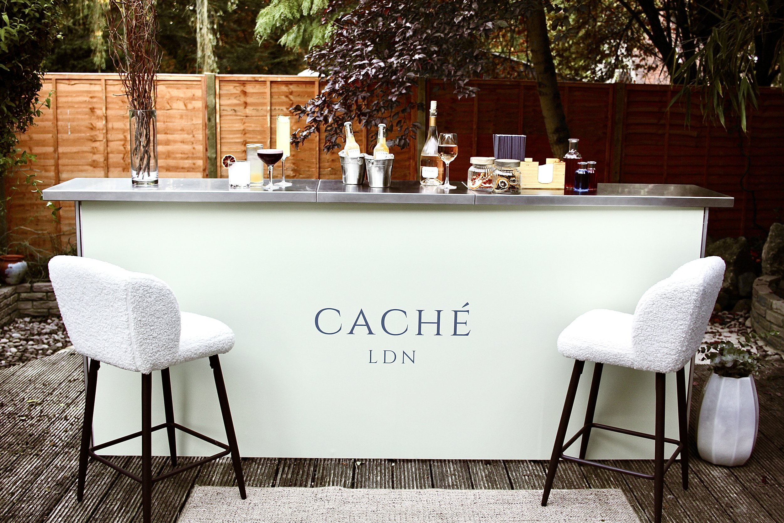 An outdoor home bar set on a wooden deck with white cushioned bar stools, various bottles, glasses, and barware on top, and a light-colored panel with the words 'CACHÉ LDN' in the center, with a garden and wooden fence in the background.