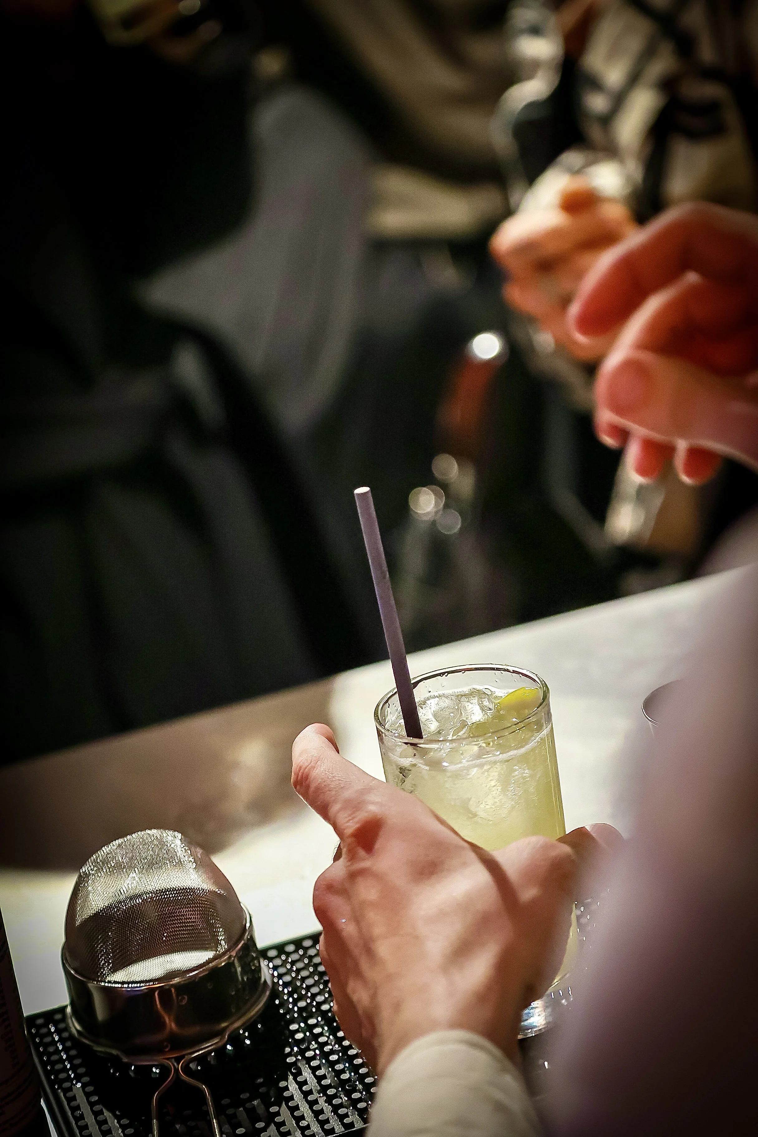 Person holding a cocktail glass with ice and lemon slice, with a bar spoon inside, on a bar counter with a metal strainer and bar tools nearby.