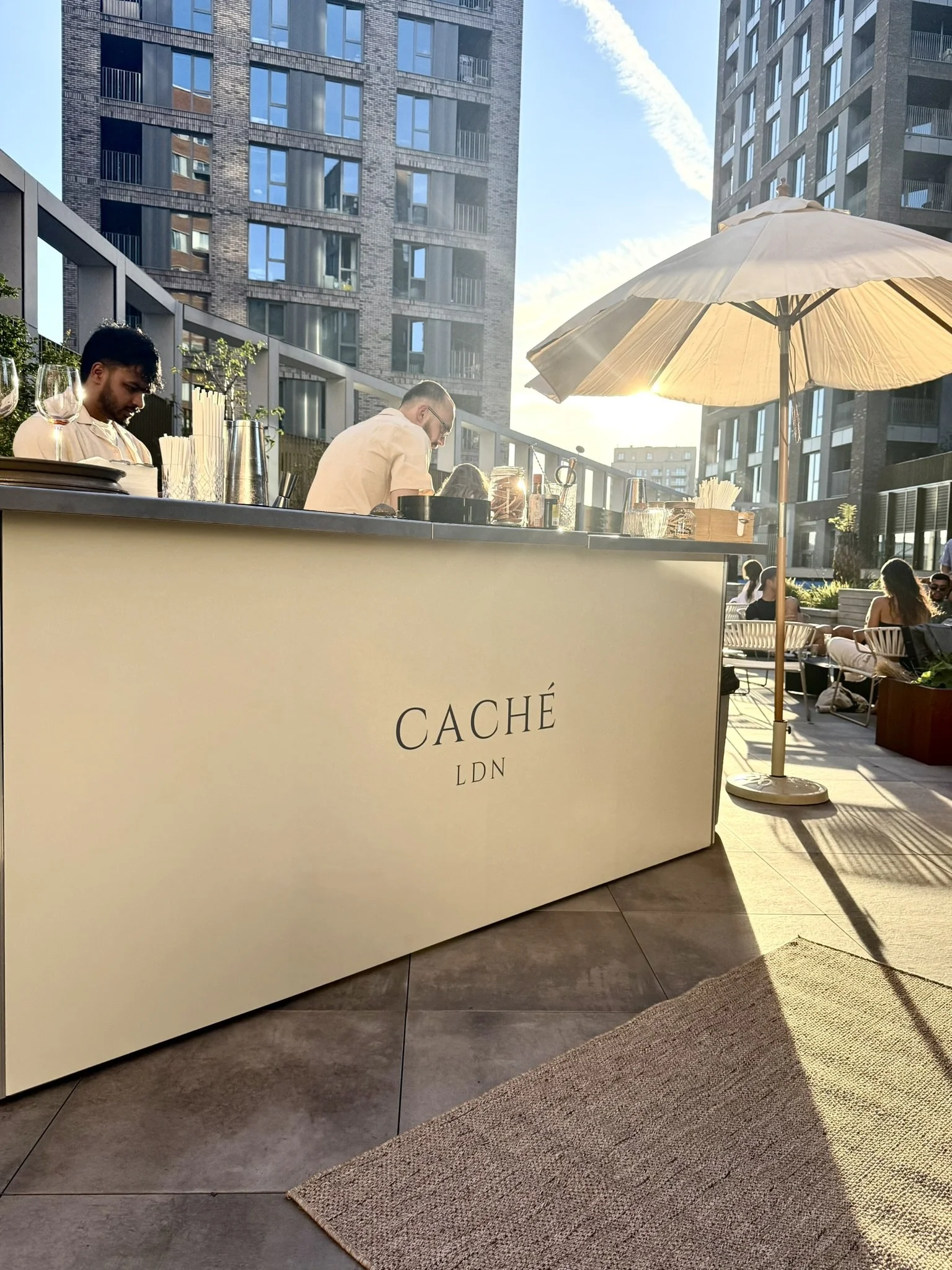 Outdoor bar area with a large umbrella, modern high-rise buildings in the background, and people sitting and enjoying the sunlight.