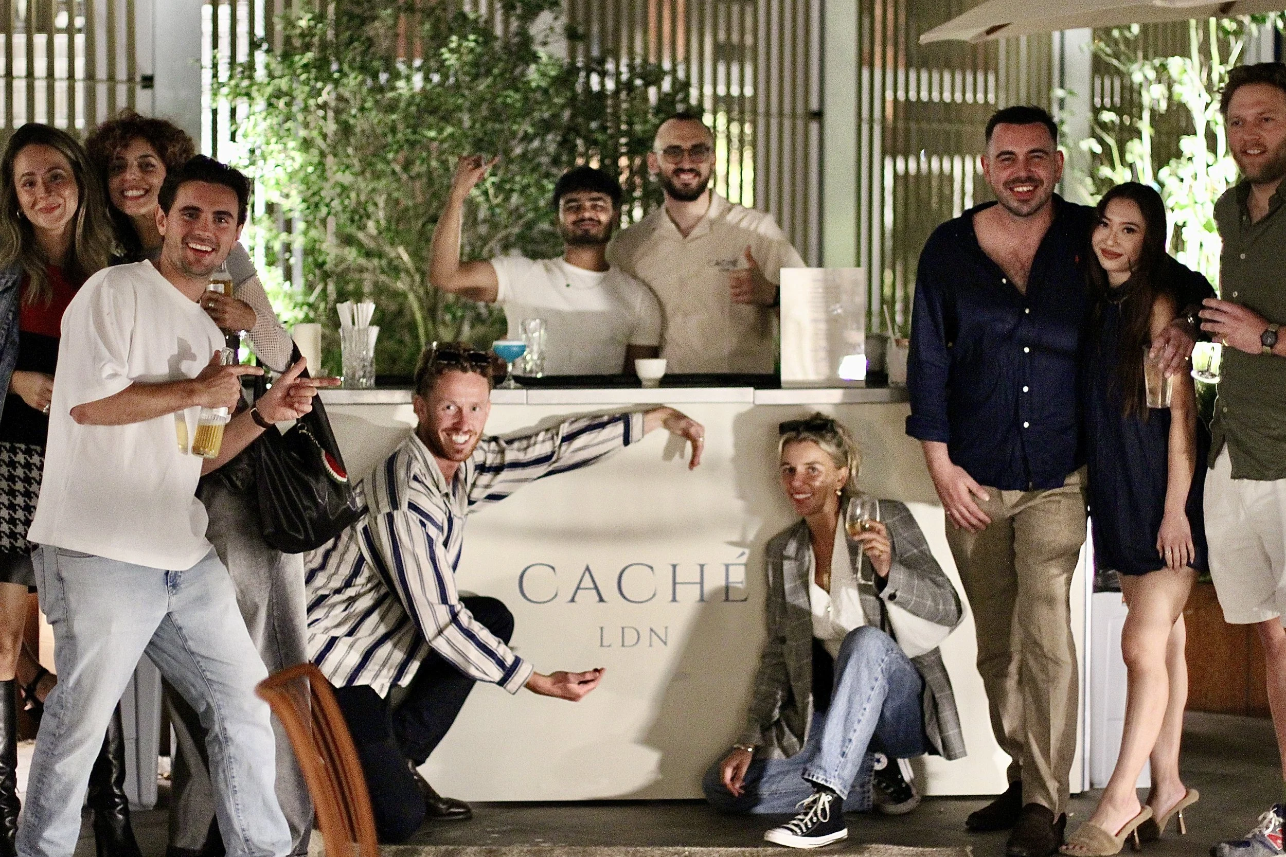 Group of people enjoying drinks at a bar, smiling and posing for the photo, with greenery in the background.