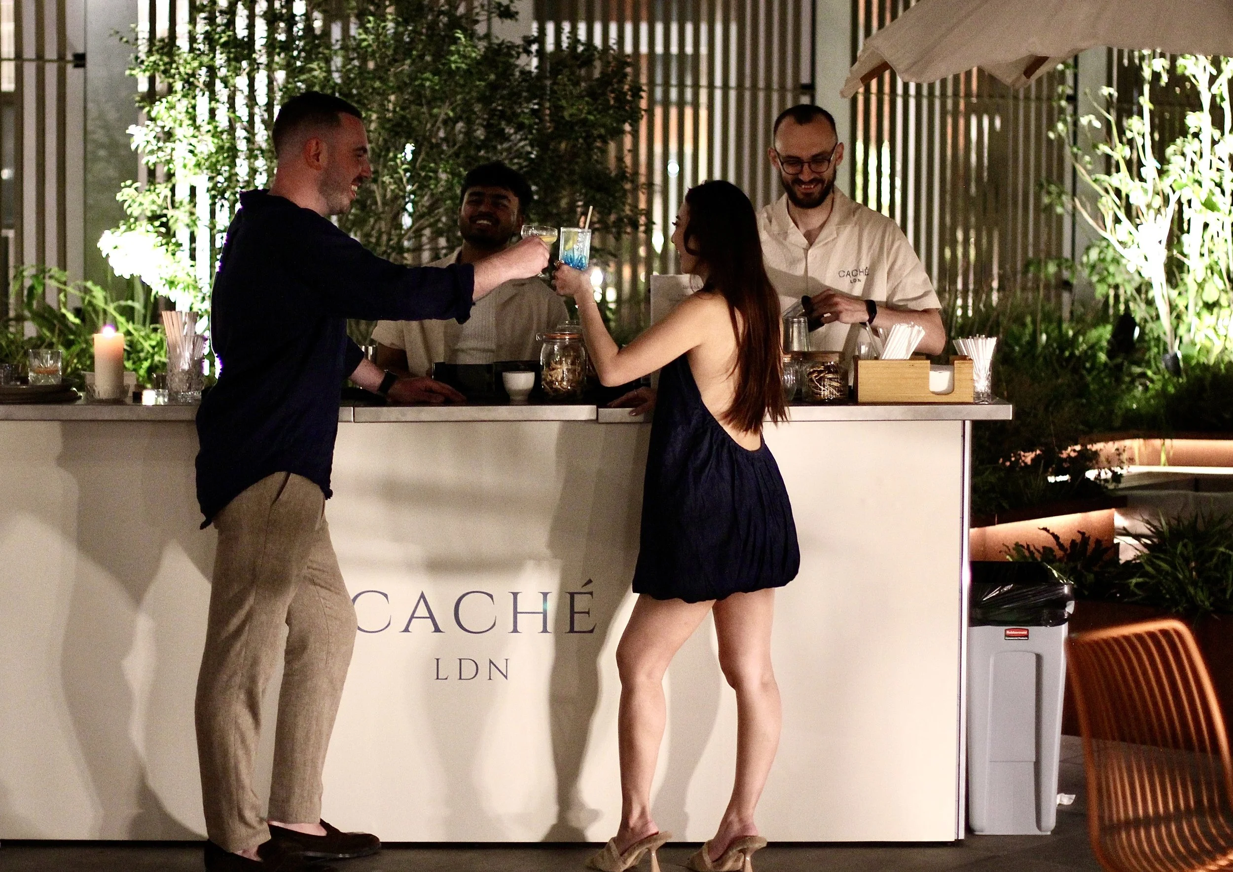 People at an outdoor bar, raising glasses for a toast, with a woman in a short, dark dress and heels, and three men behind the bar smiling, at night with lush greenery and decorative lighting.
