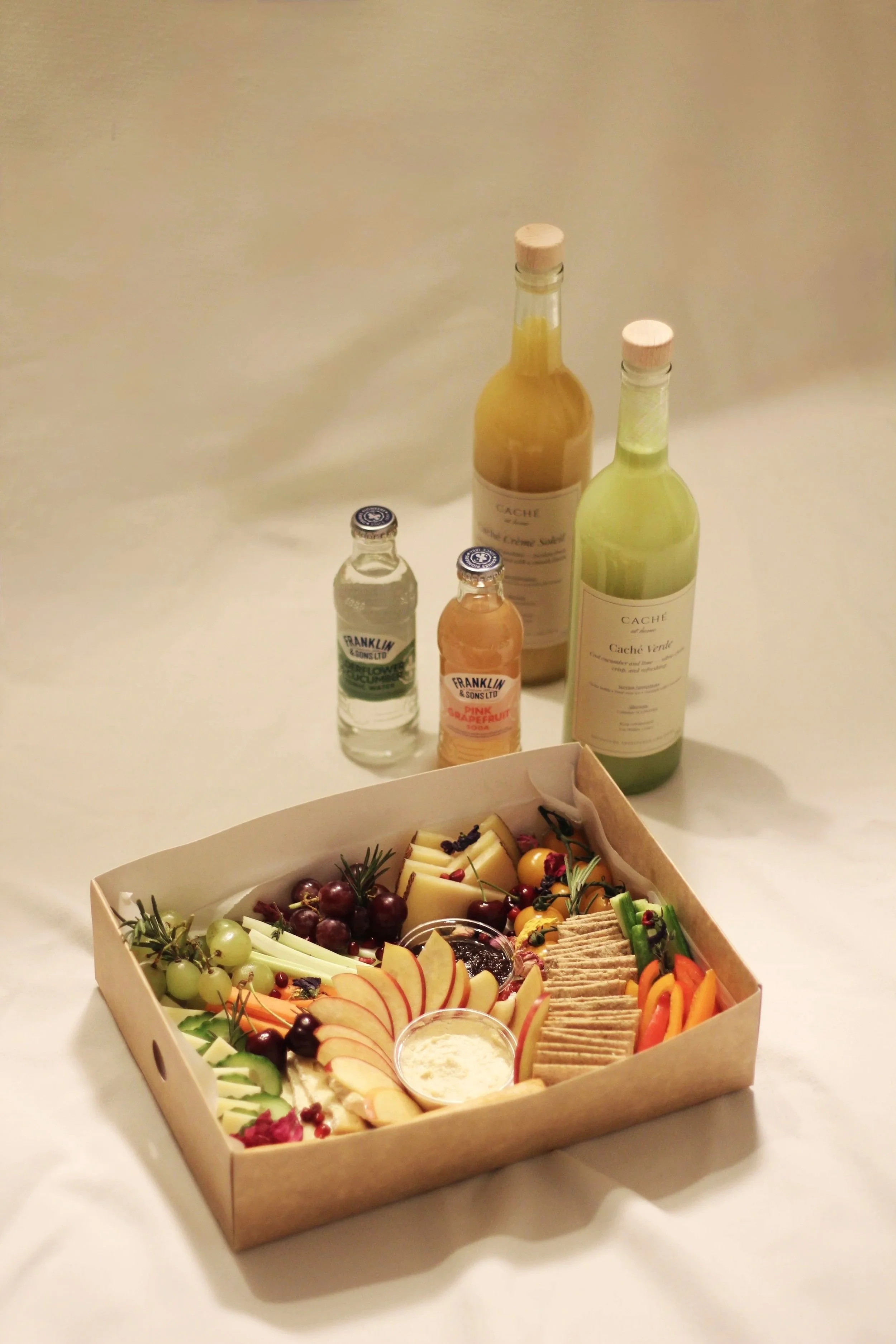 A cheese and fruit platter with grapes, apple slices, and crackers, alongside bottles of wine and flavored drinks on a white surface.
