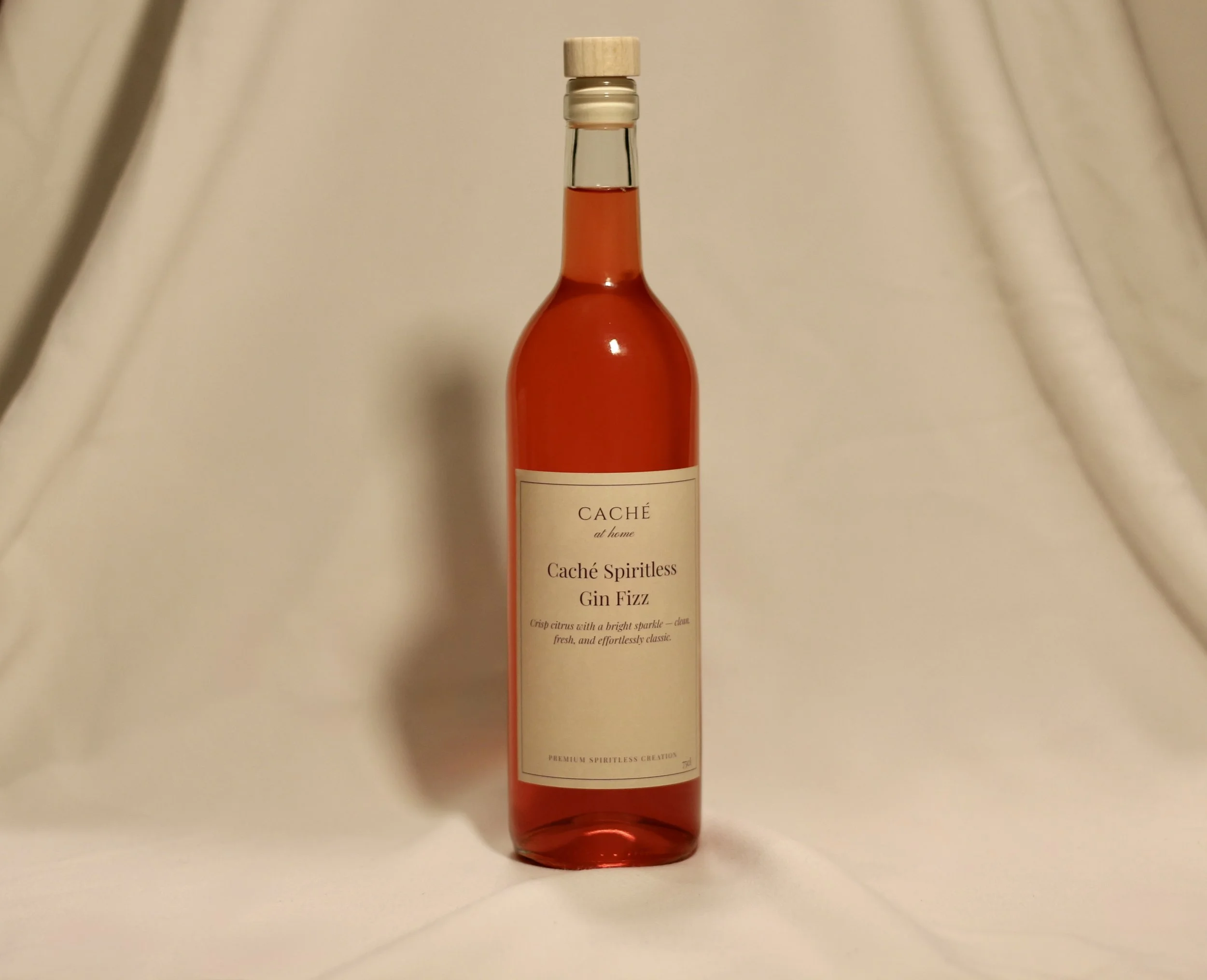 A bottle of Caché Spiritless Gin Fizz with a light-colored label and a wooden cap, standing on a white surface with a plain background.