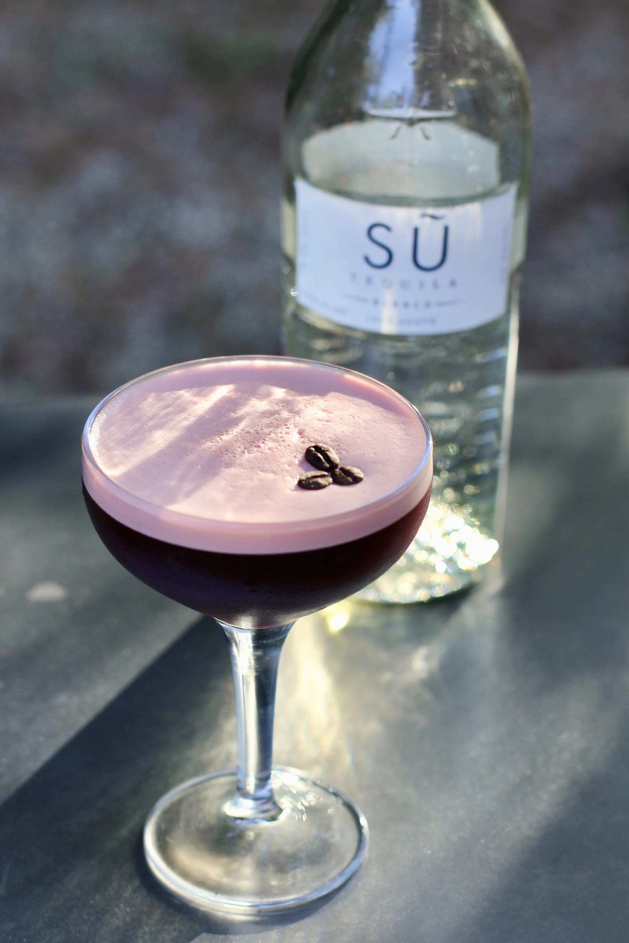 A pink cocktail with three coffee beans garnished on top, positioned in the foreground, with a bottle of Su Trefosa mineral water in the background.
