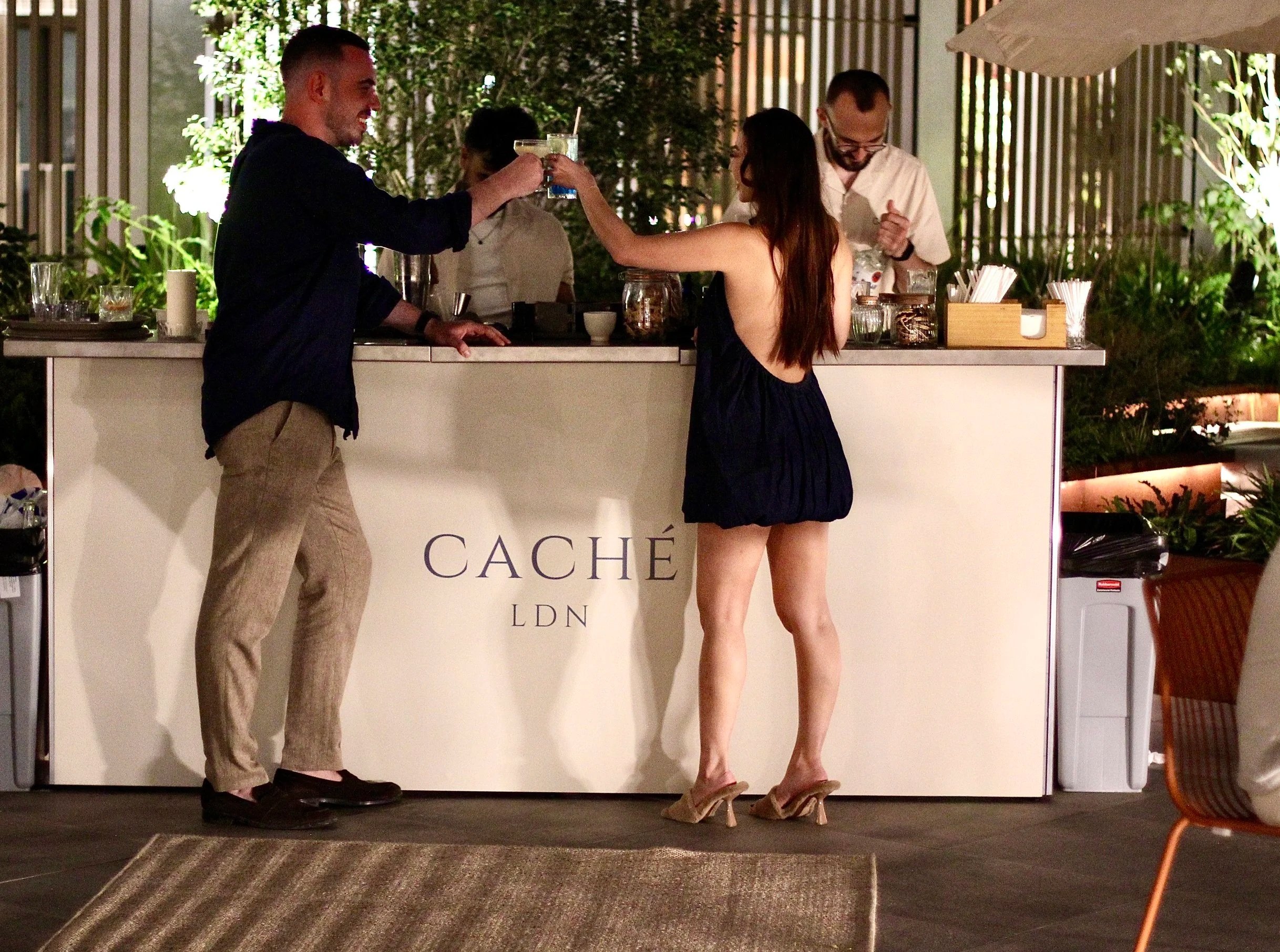 Nighttime outdoor bar scene with three people, a woman in a short dress and heels, a man in a dark shirt and beige pants, and two bartenders behind the bar, one in a white shirt, all at a bar labeled 'CACHÉ LDN' with greenery and lighting in the background.