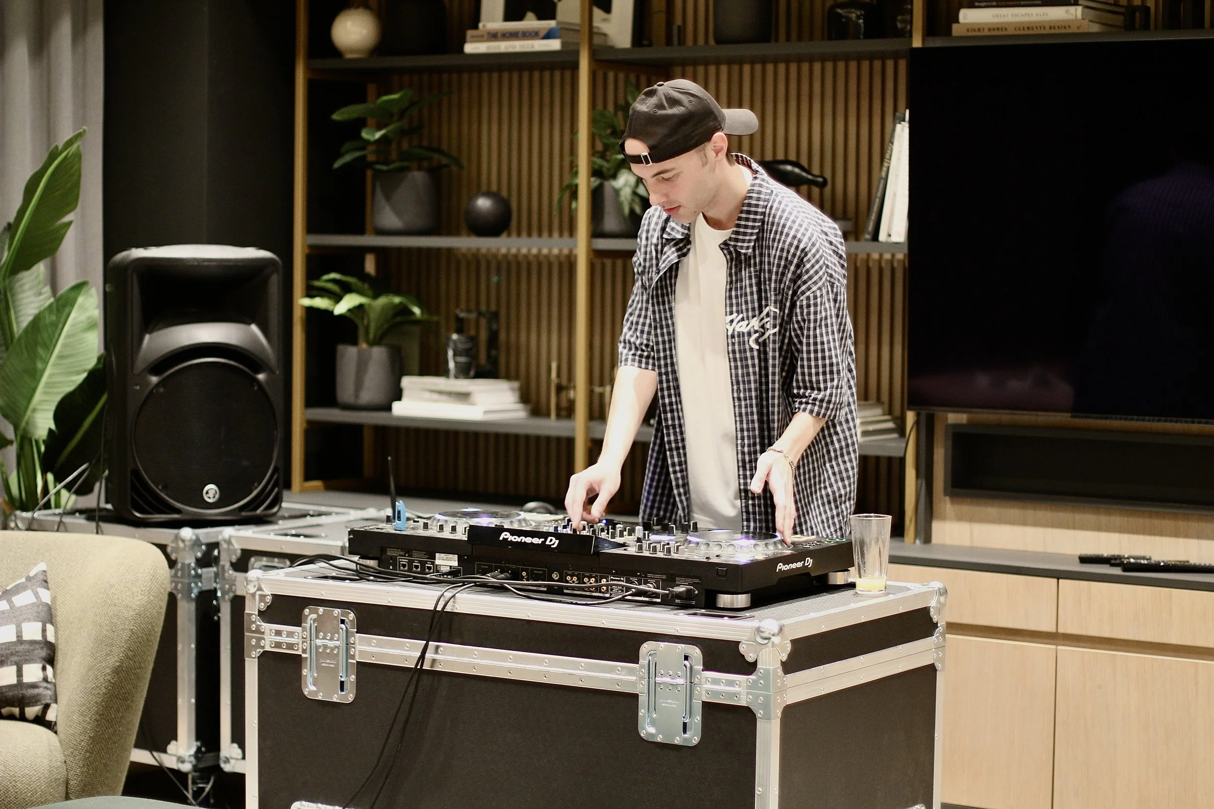 A DJ wearing a backward baseball cap and plaid shirt is mixing music on a Pioneer DJ controller in a modern living room or lounge area, with a large speaker, plants, and a bookshelf in the background.