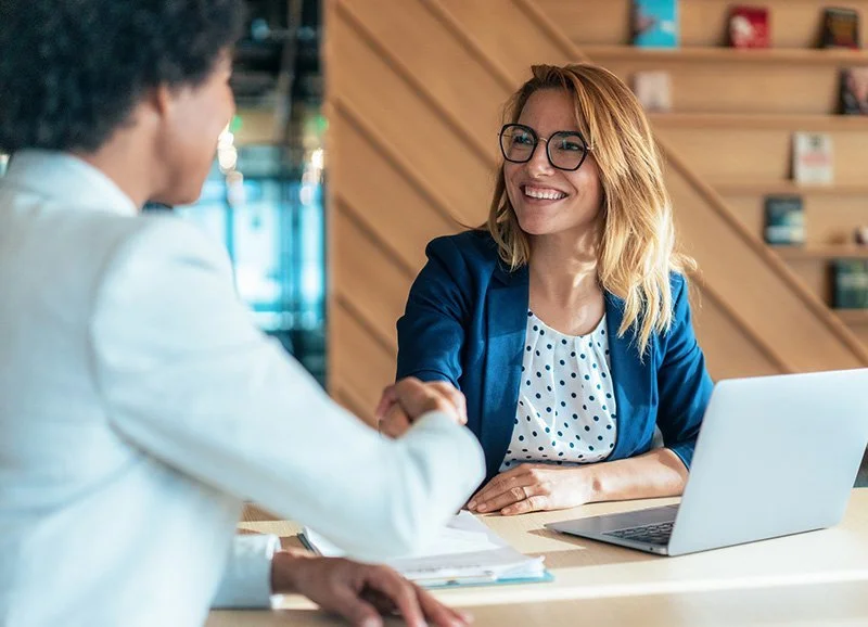 A woman in a blue blazer and polka dot blouse smiling and shaking hands with a person in a white blazer across a table in a modern office or coffee shop setting, with a laptop and papers on the table.