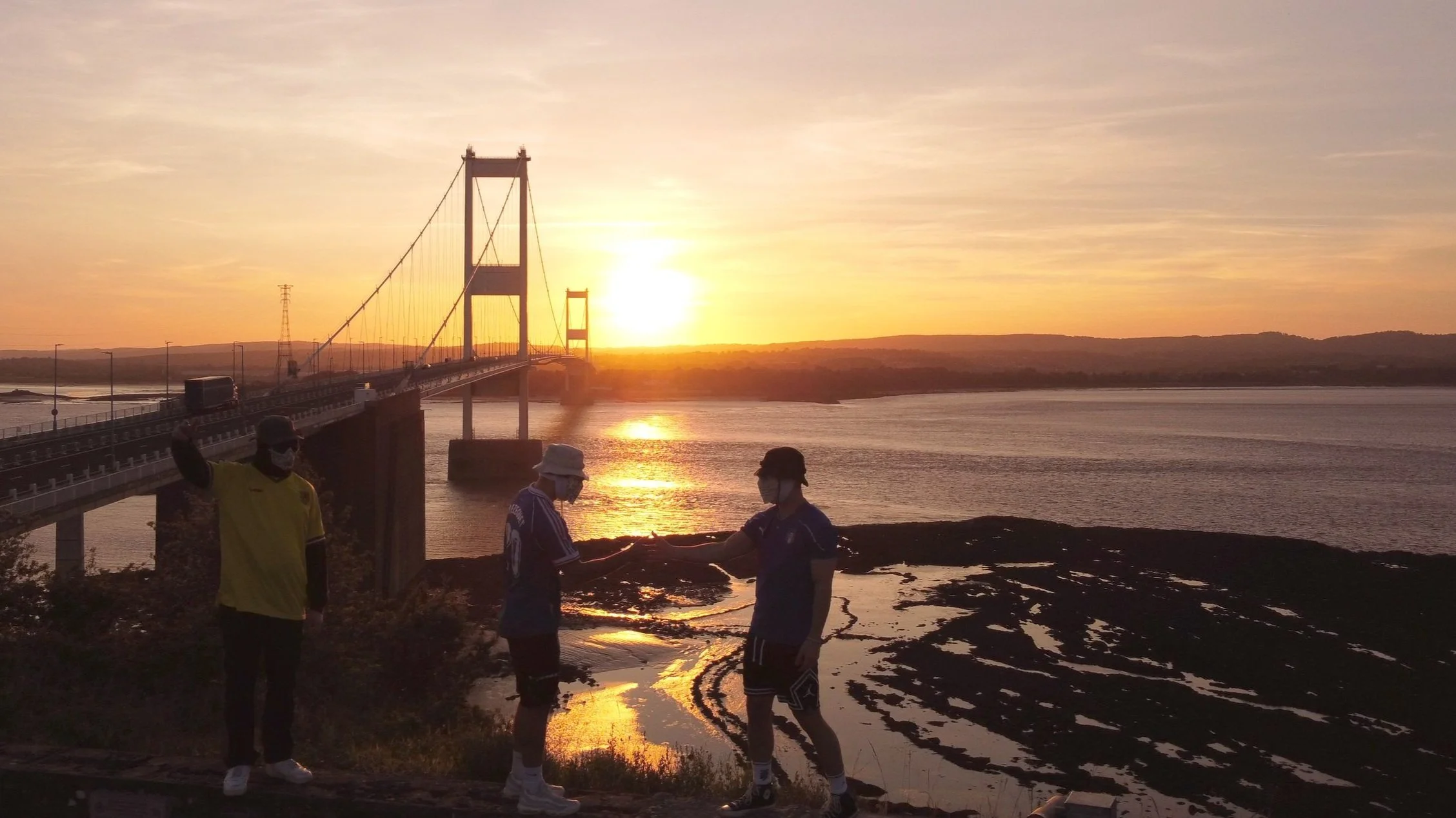 Sunset over a body of water with a bridge in the background and three people in the foreground, two of whom are exchanging something while wearing masks.