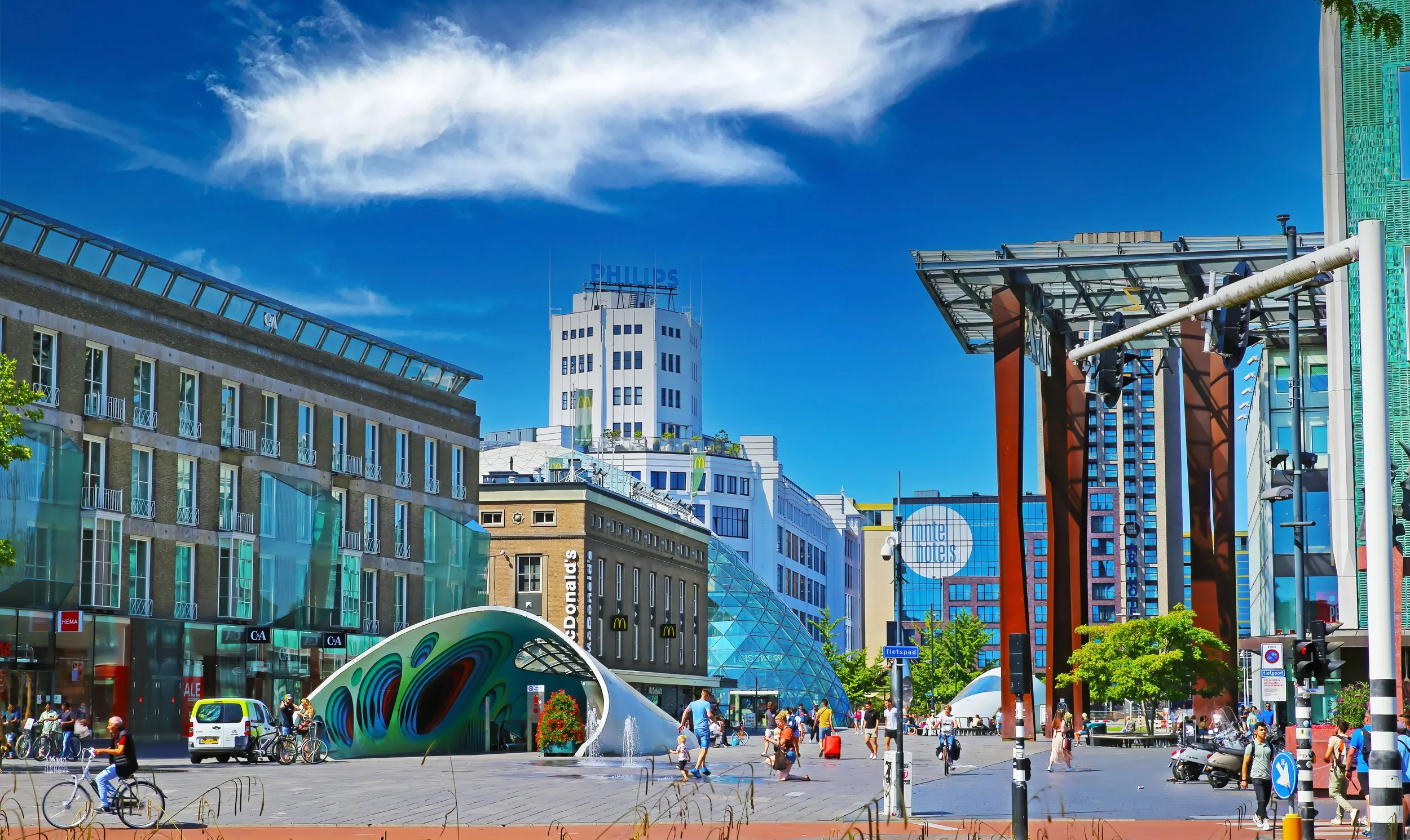 Urban cityscape with modern buildings, a glass-domed entrance, people walking, biking, and a fountain, under a bright blue sky with clouds.