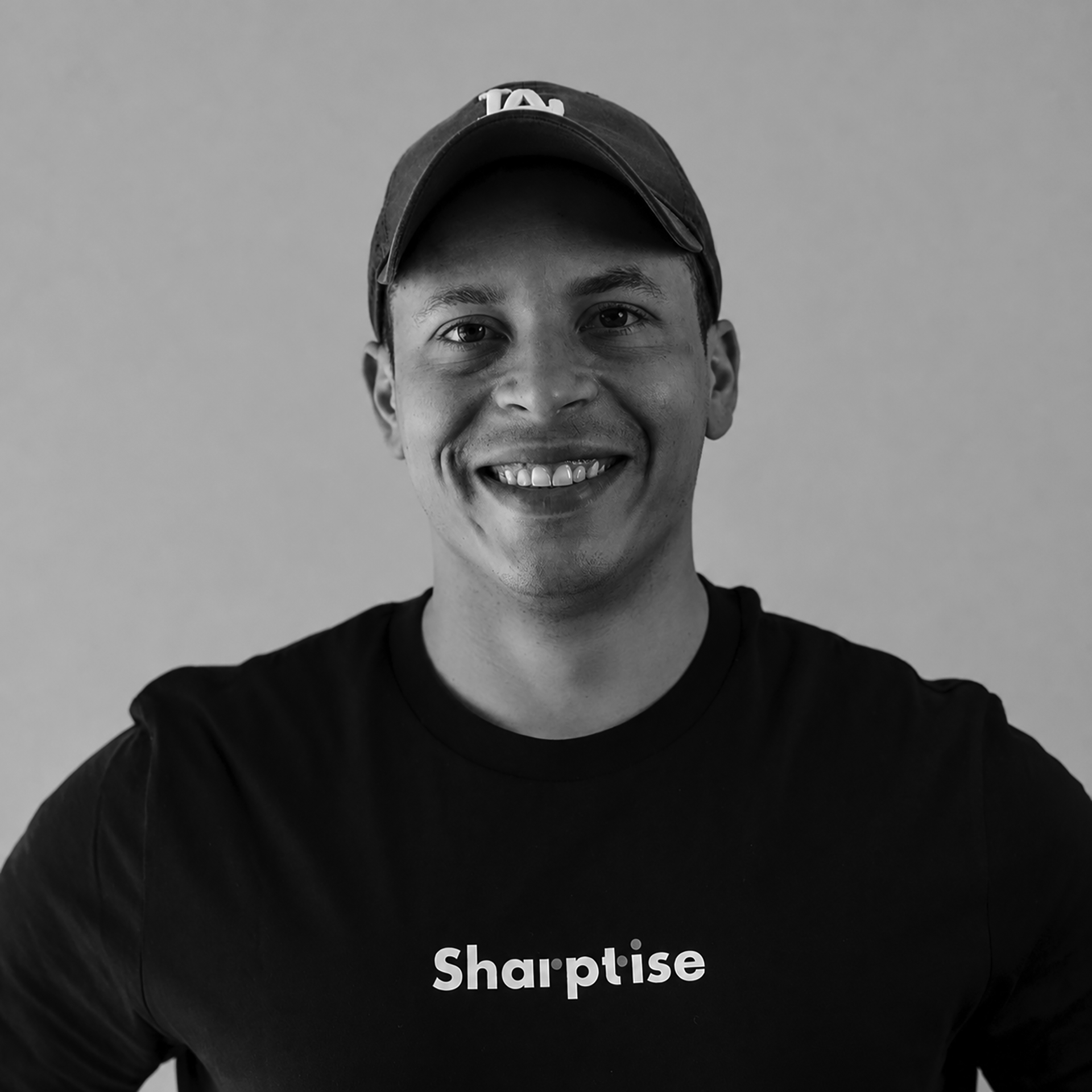 Close-up of a young man wearing a baseball cap and a black t-shirt with the text 'Sharptise' printed on it, smiling and looking at the camera against a plain background.