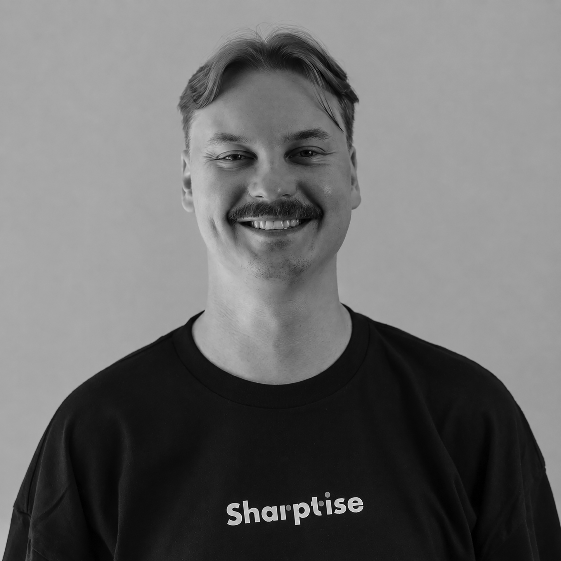 Close-up black and white photo of a smiling man with short hair, mustache, and wearing a black t-shirt with white text that says 'Sharetise'.