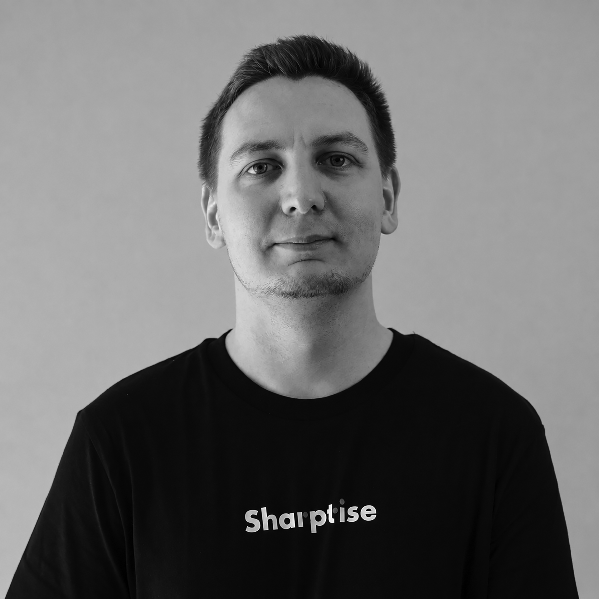 Black and white portrait of a young man with short dark hair, light skin, wearing a black shirt with the word 'sharpise' printed on it.