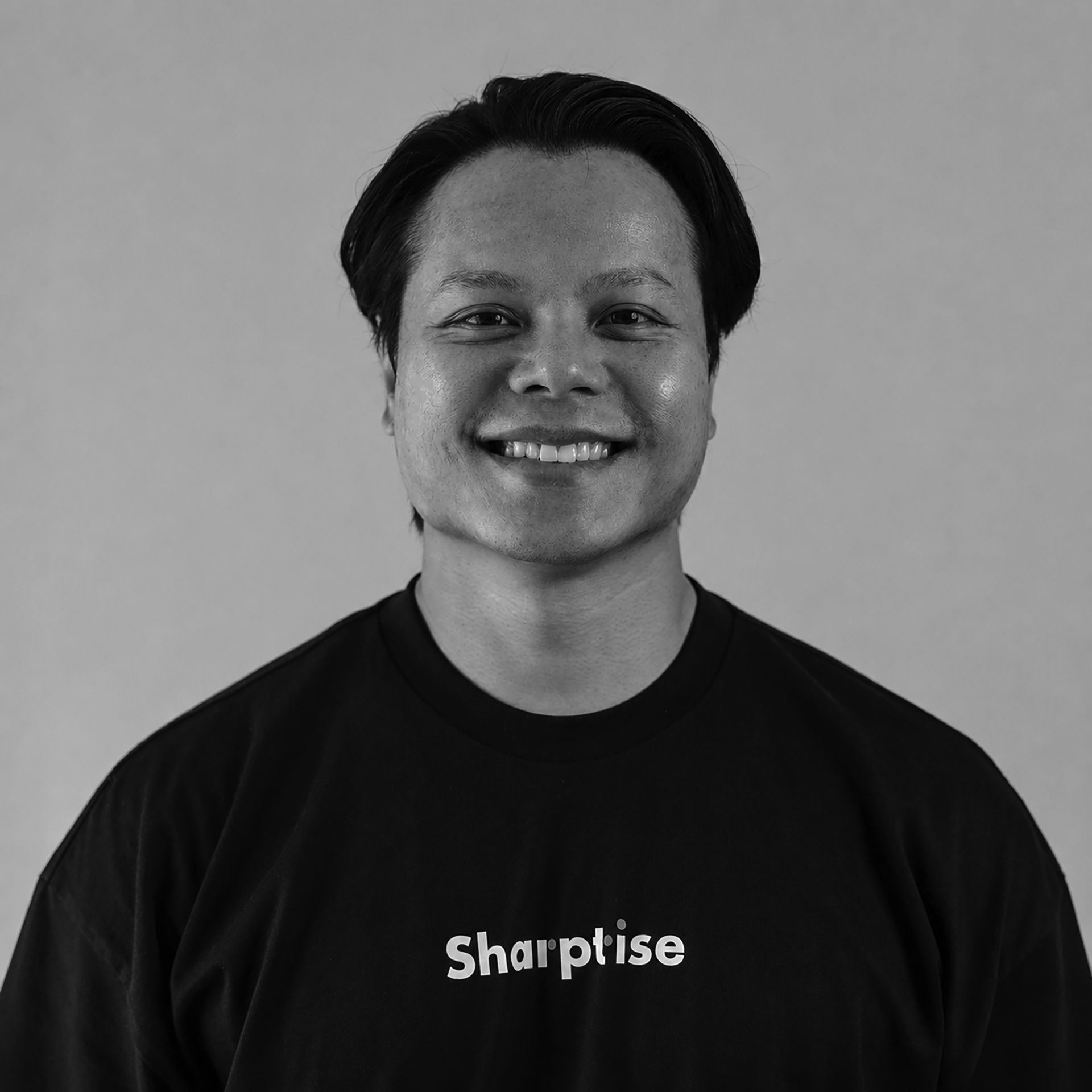 Close-up of a person with short dark hair smiling, wearing a black shirt with the word 'Sharptise' written on it.