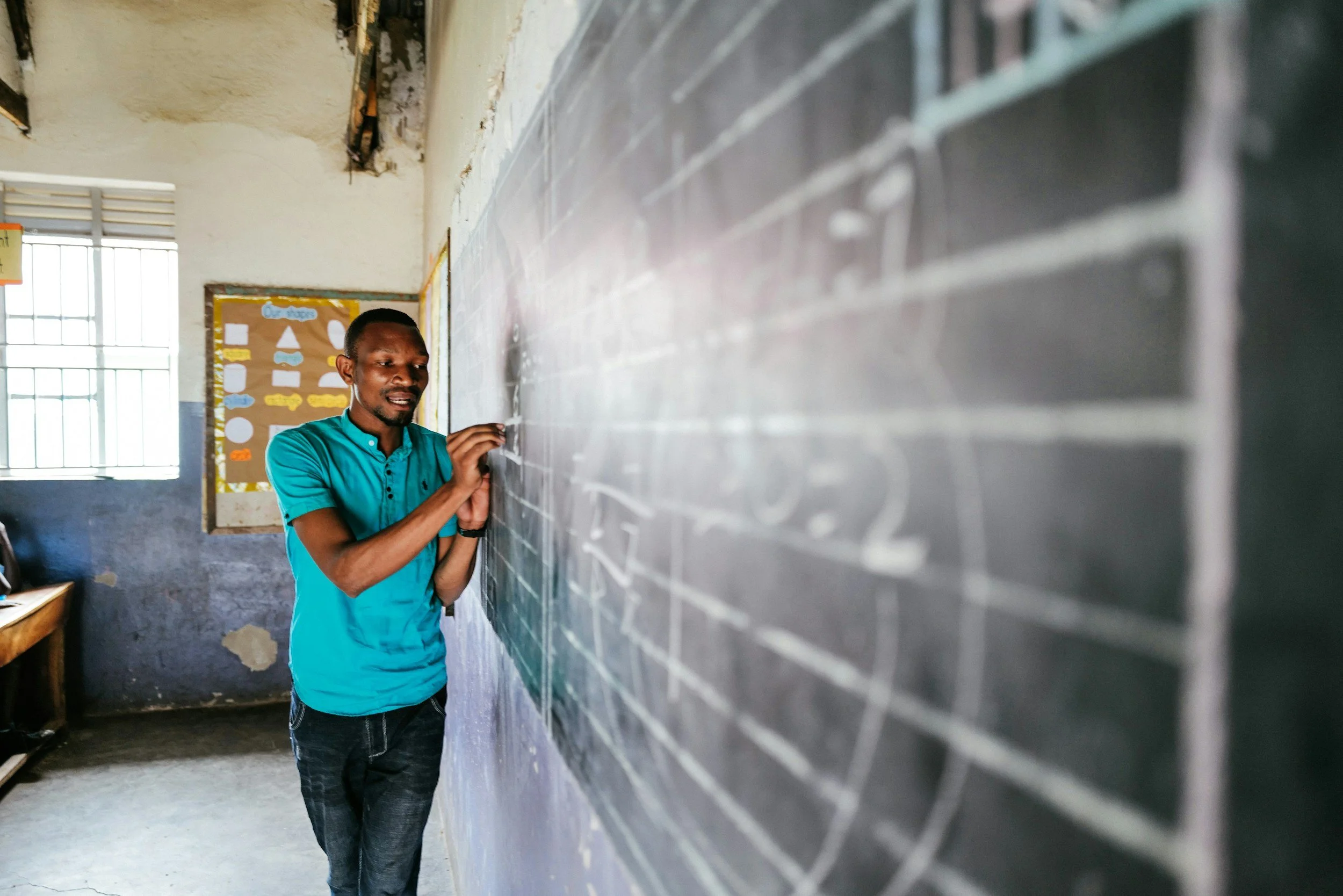 A man writing on a chalkboard in a classroom with a bulletin board and window in the background.