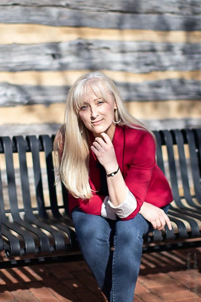 Confident female caregiver with long blonde hair, seated outdoors in casual professional attire—red blazer and jeans—on a black bench, gently smiling with hand on chin against a warm wooden backdrop