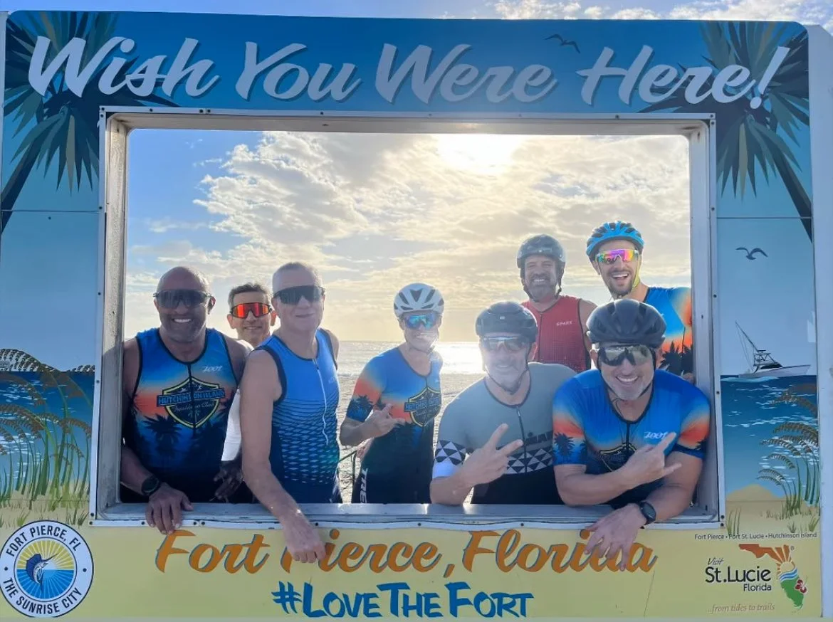 Group of eight cyclists posing for a photo at the beach behind an arch with 'Wish You Were Here!' written on top, with the sun setting over the ocean in the background. They are wearing cycling gear and helmets, smiling and making hand gestures, with a mostly clear sky.