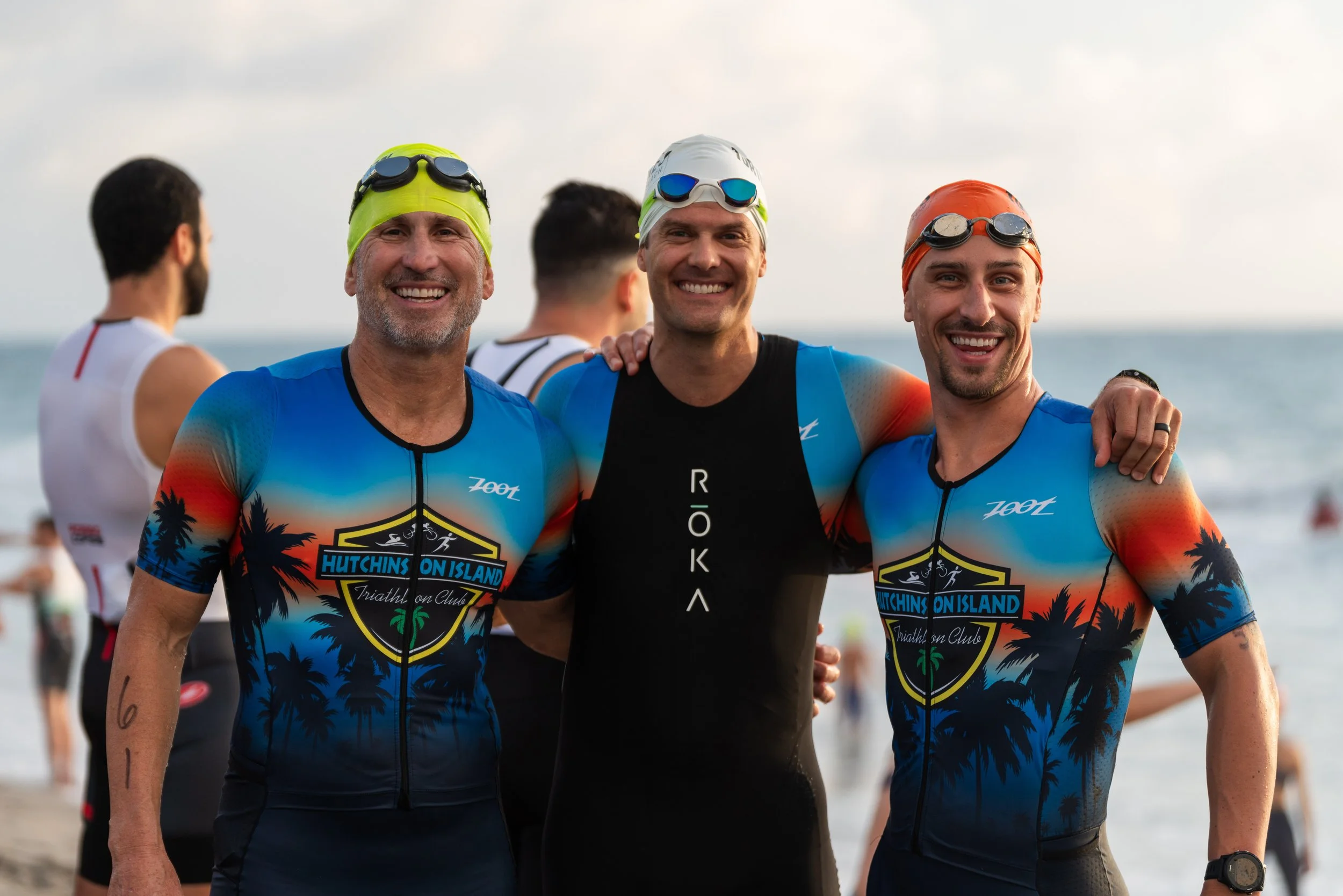Three smiling male triathletes in wetsuits and swim caps posing together at the beach, with the ocean in the background.