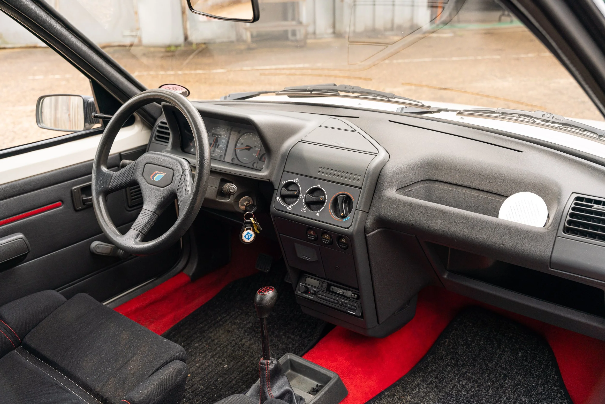Interior of a vintage car with black dashboard, red and black carpet, manual gear shift, three circular control knobs on the center console, and a steering wheel with a logo.