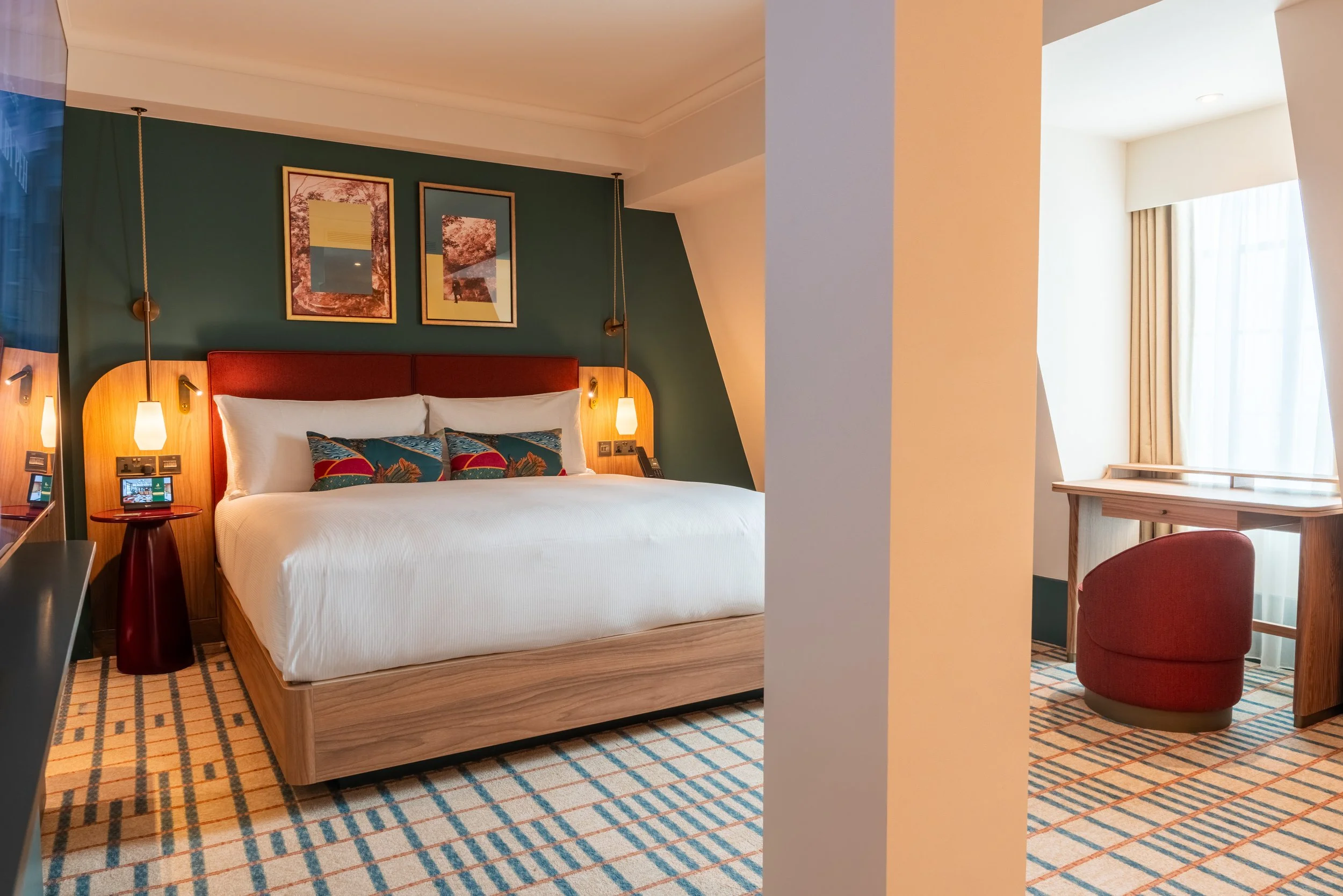 A hotel room with a bed, two framed pictures above it, a green accent wall, hanging wall lamps, a small round side table, and a window with beige curtains. There is also a desk with a red chair near the window.