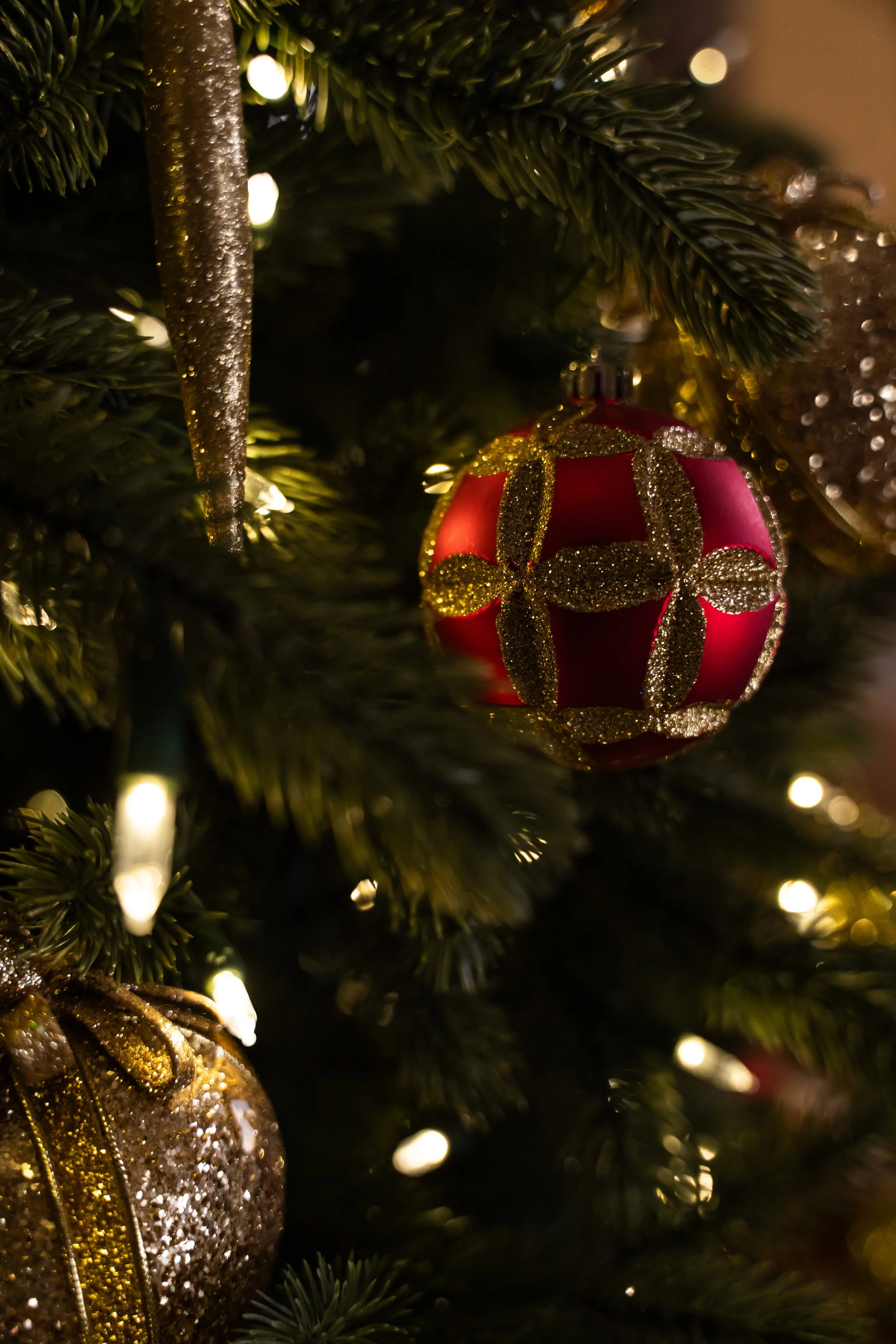 Close-up of a Christmas tree with ornaments, including a red ball with gold glitter and ribbon pattern, surrounded by pine branches and small lights.