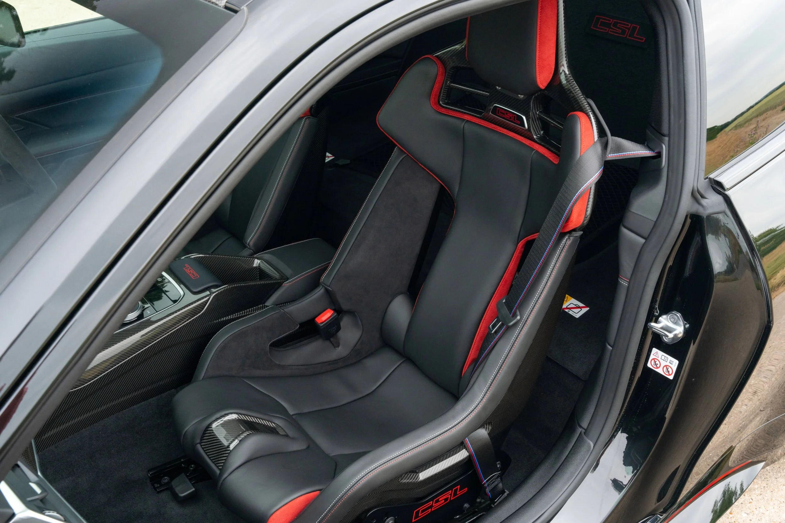 Inside view of a luxury sports car showing black leather racing seats with red stitching, carbon fiber accents, and 'CSL' branding.