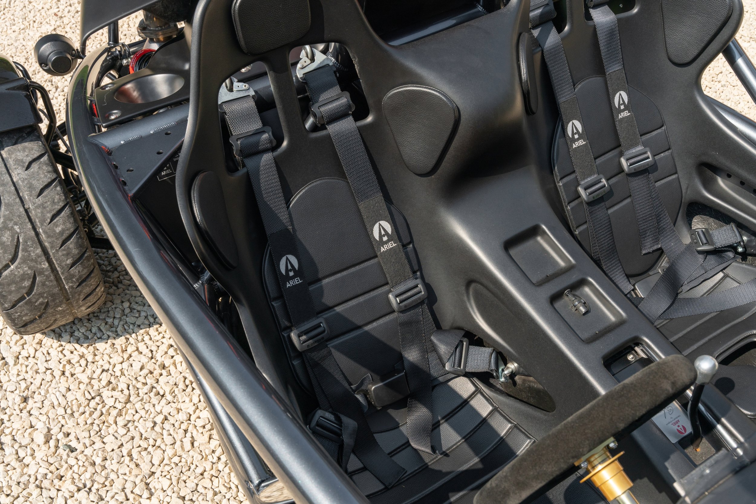 Two black racing seats inside a lightweight vehicle with harness belts, surrounded by a black metal frame, on a gravel surface.
