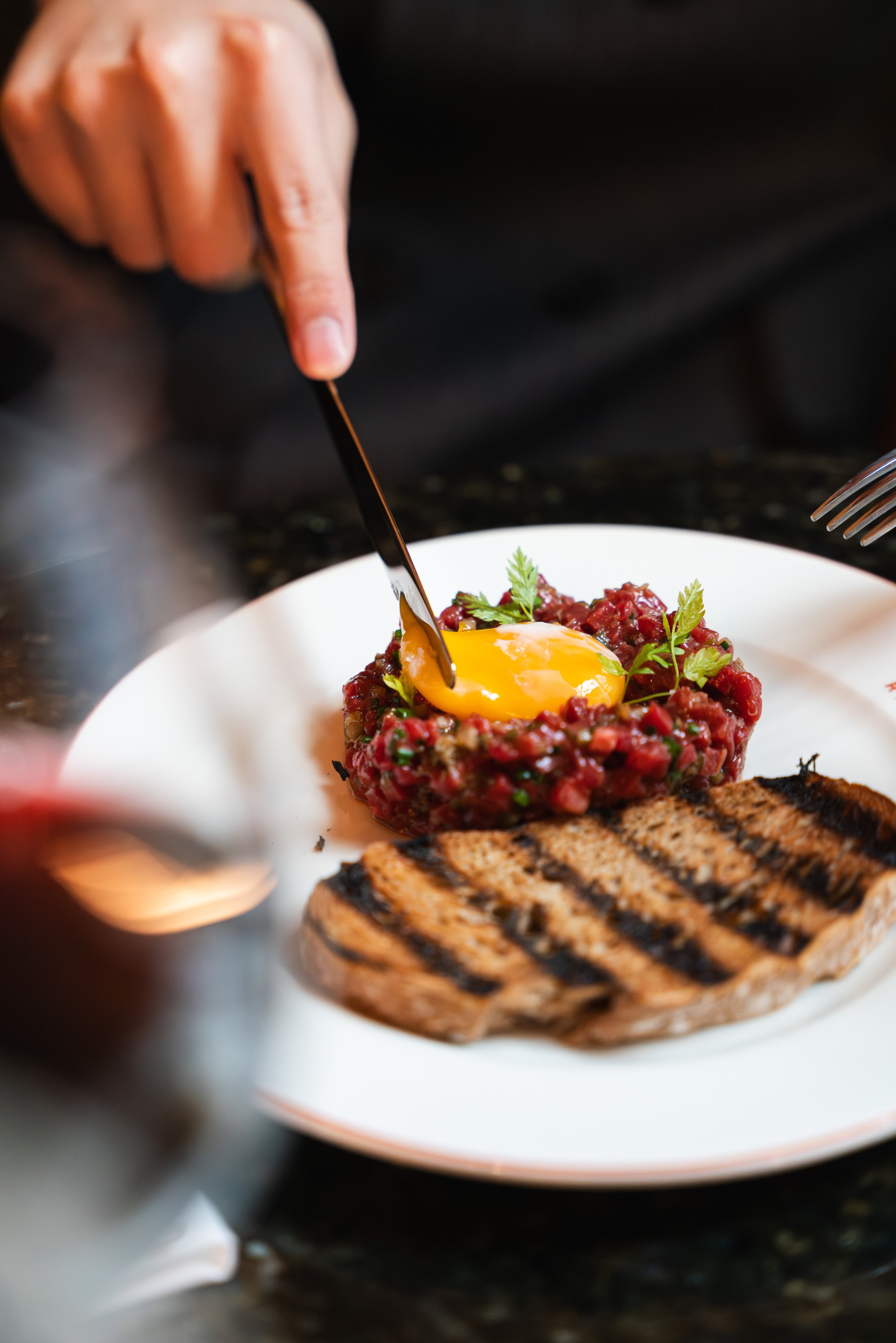 A close-up of a raw steak tartare topped with a raw egg yolk, garnished with fresh herbs, served on a white plate with a grilled piece of bread nearby.