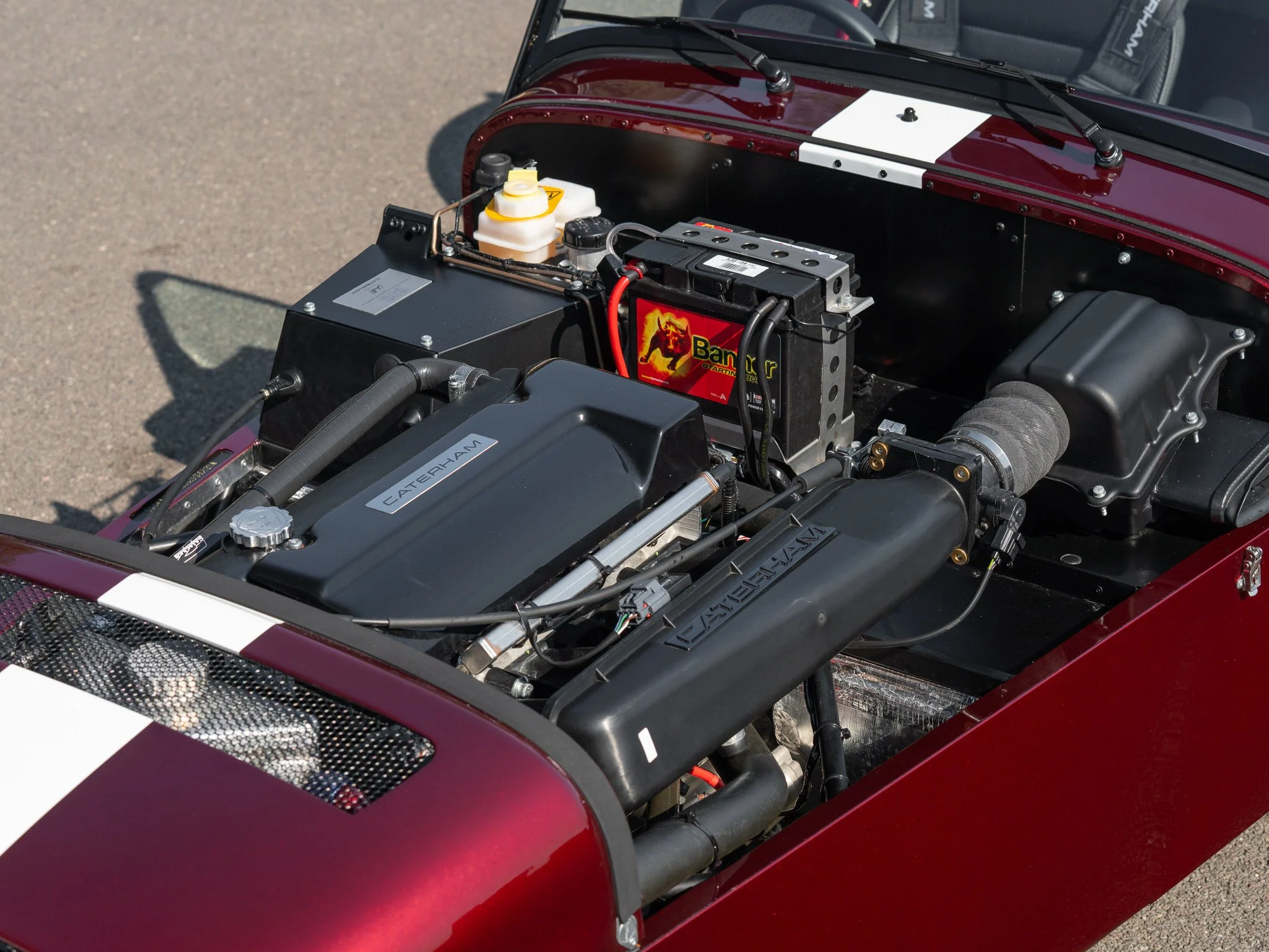 Close-up of a race car engine with a Caterham branding, a large battery labeled 'Balltr,' and other engine components in a red and black chassis.