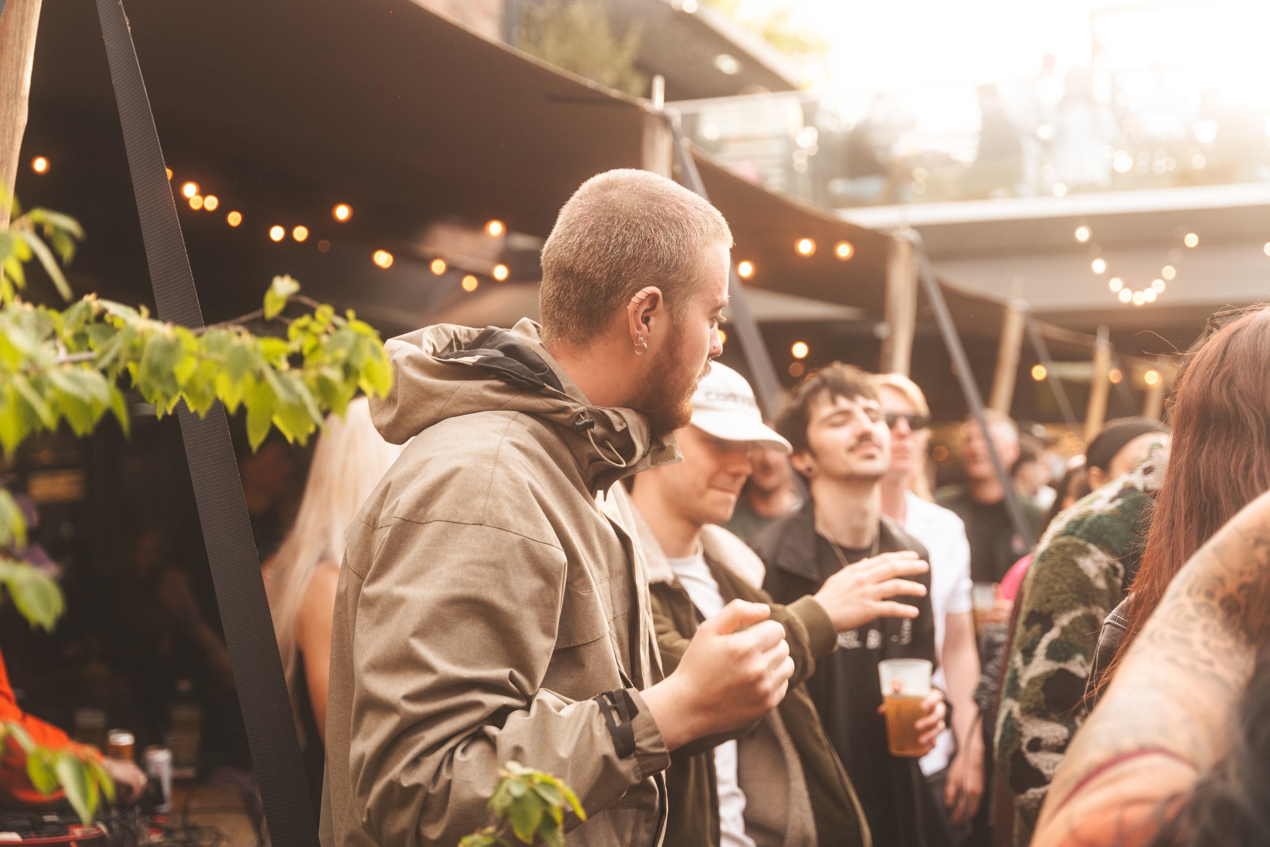 A group of people at an outdoor event during sunset, with string lights overhead, some holding drinks, and one person dancing or swaying.