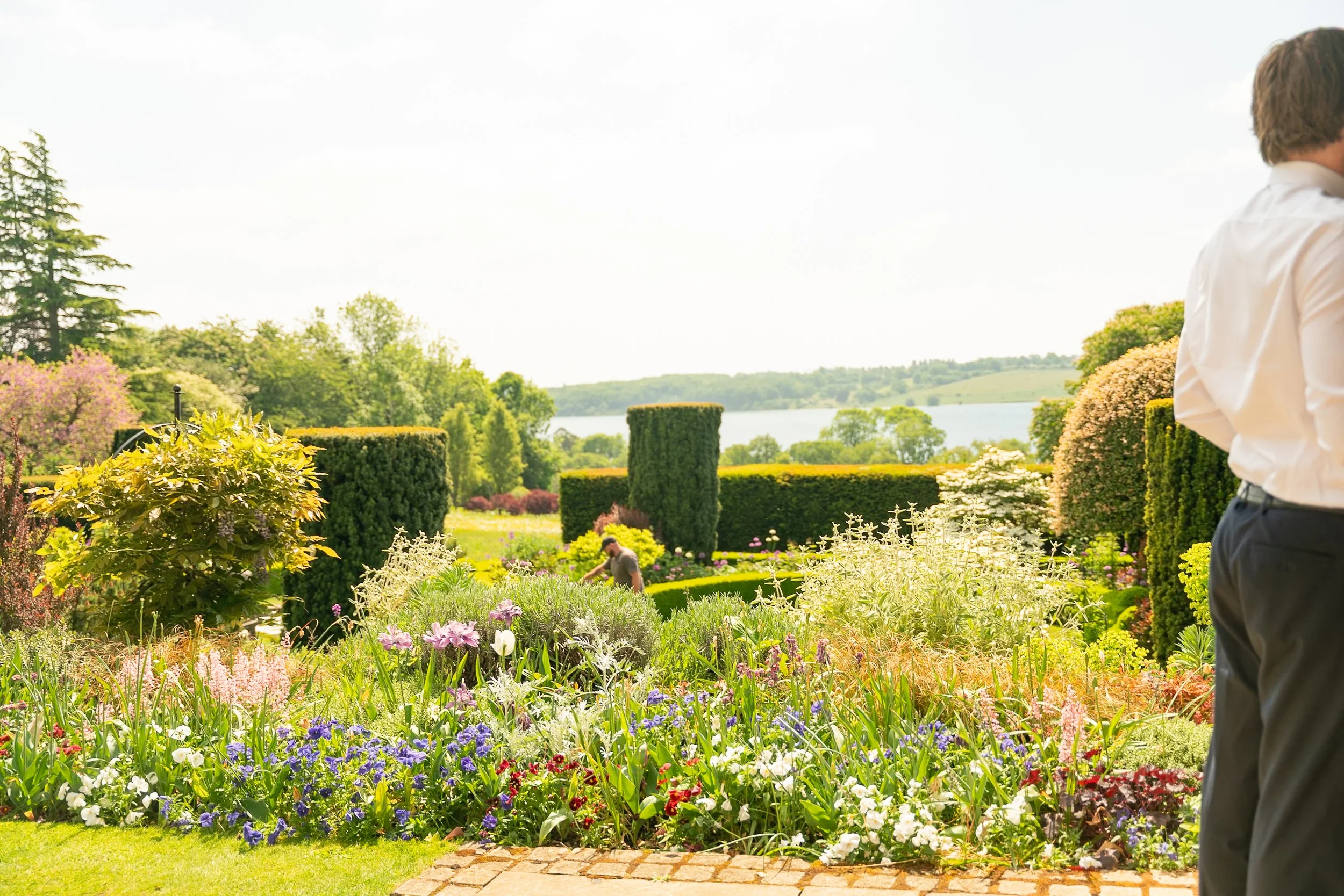 A vibrant garden with colorful flowers, trimmed shrubbery, and a scenic water view in the background on a sunny day.