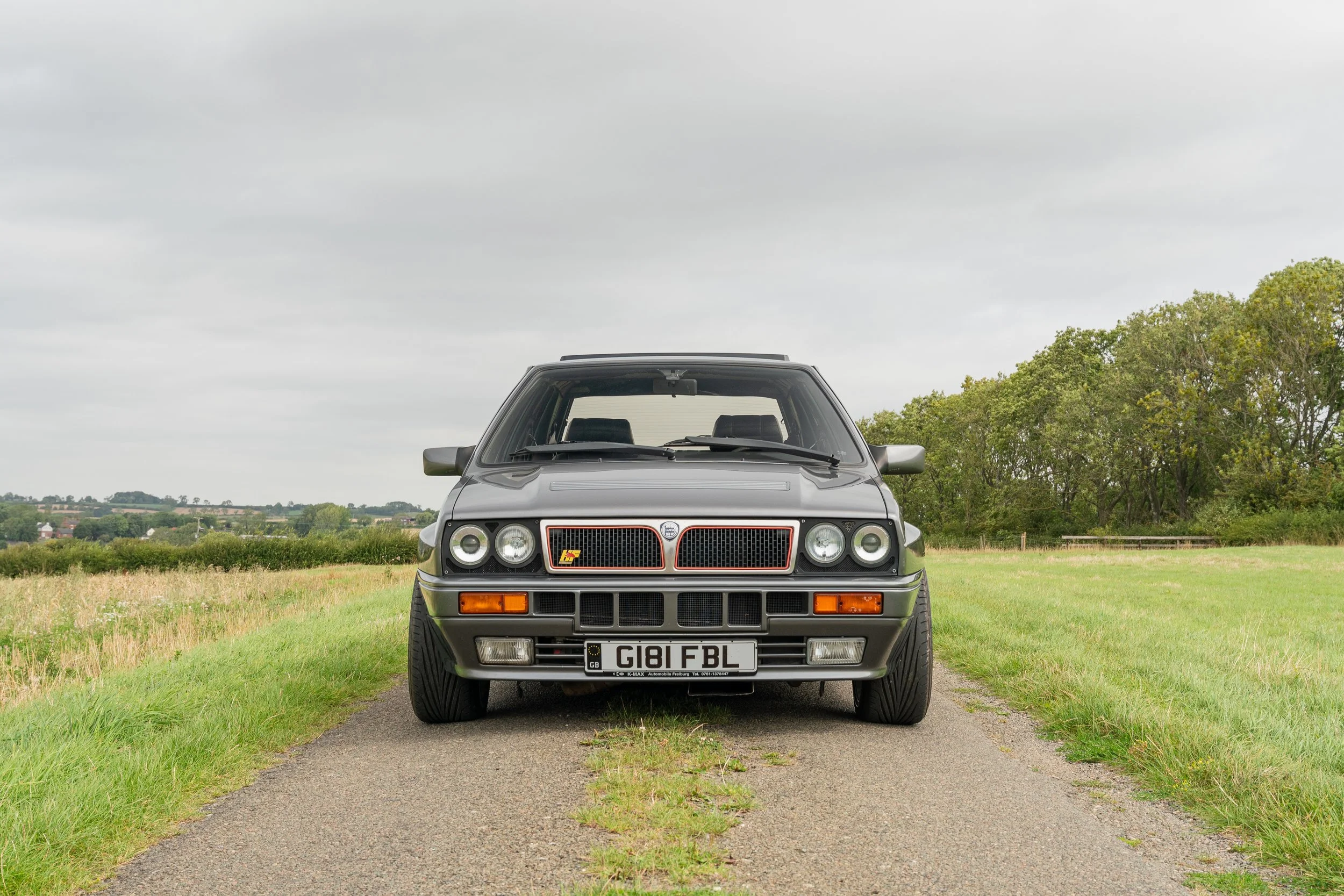 Front view of a classic black Lancia Delta Integrale parked on a rural gravel road with green fields and trees in the background.