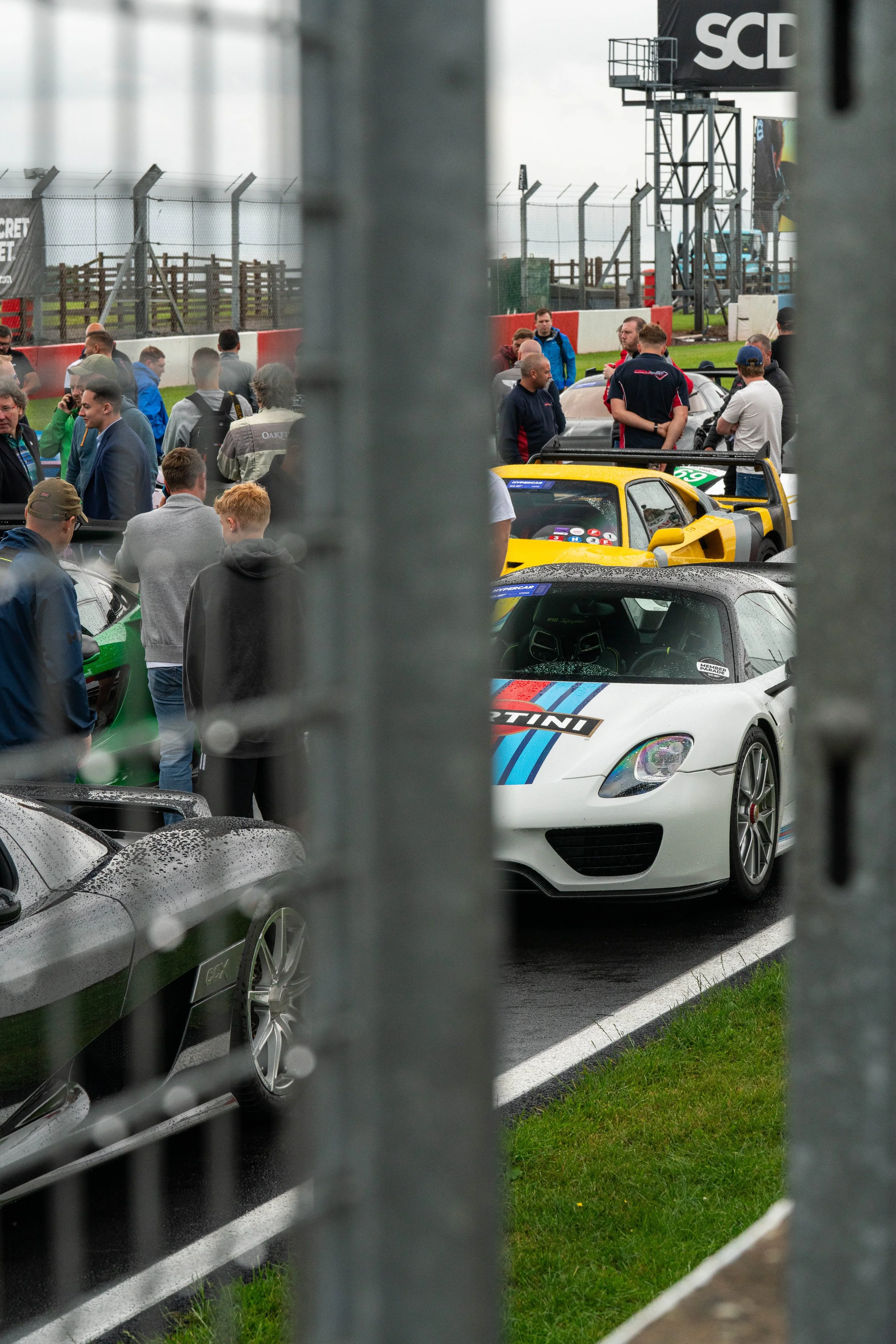 Race cars and spectators behind a fence at a motorsport event during rainy weather.