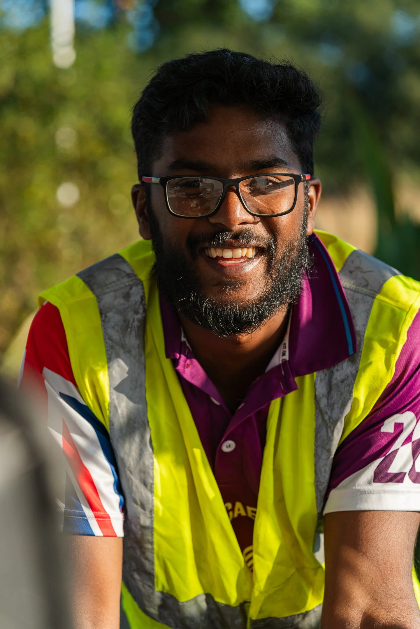A smiling man with glasses, beard, and dark hair outdoors, wearing a yellow safety vest over a purple and white sports shirt.