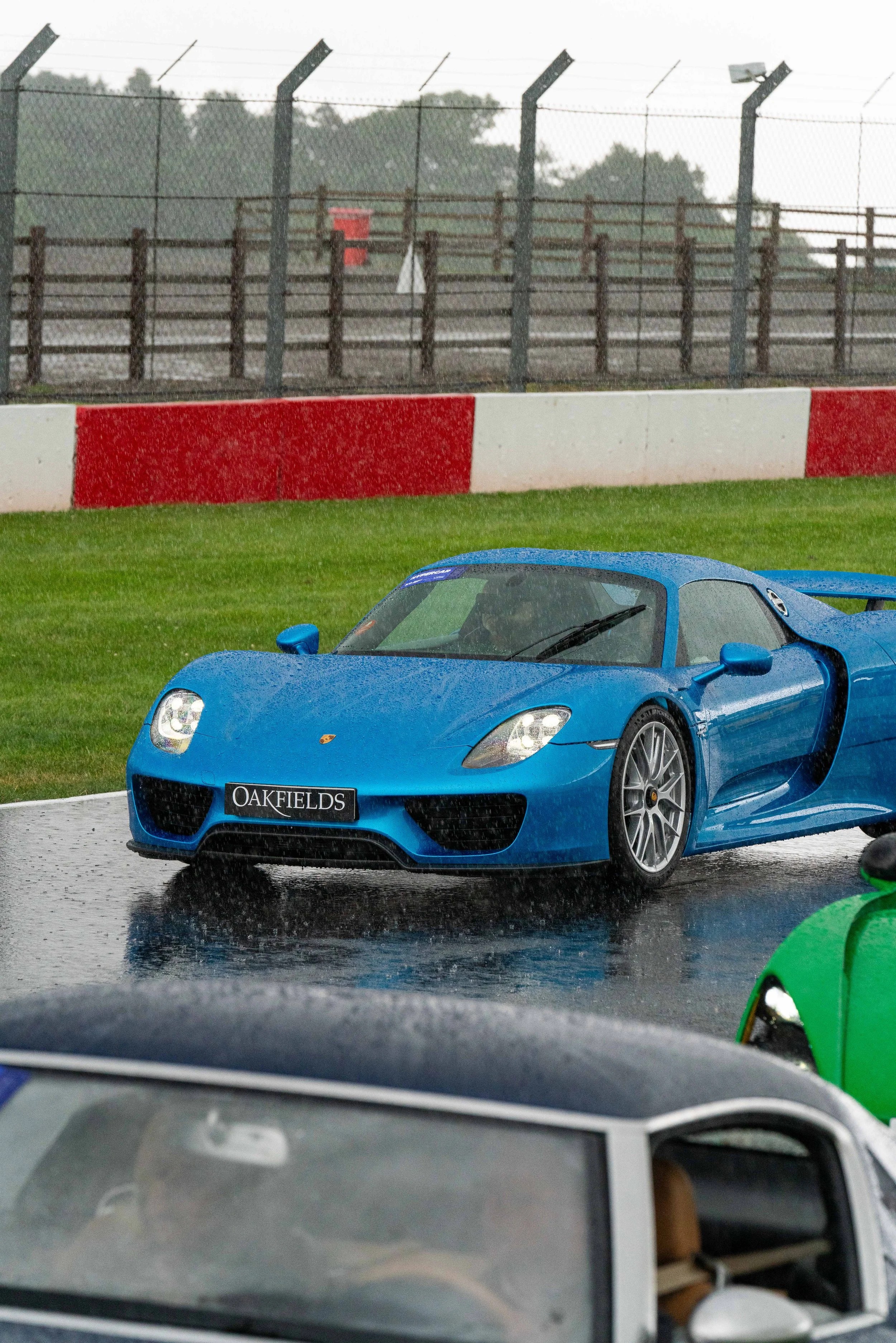 A blue sports car on a wet race track during rainy weather at a motorsport event. The car is a sleek, low-profile model with a black front grille and the word 'OAKFIELDS' on the license plate. Water droplets are visible on the car, and a green car is