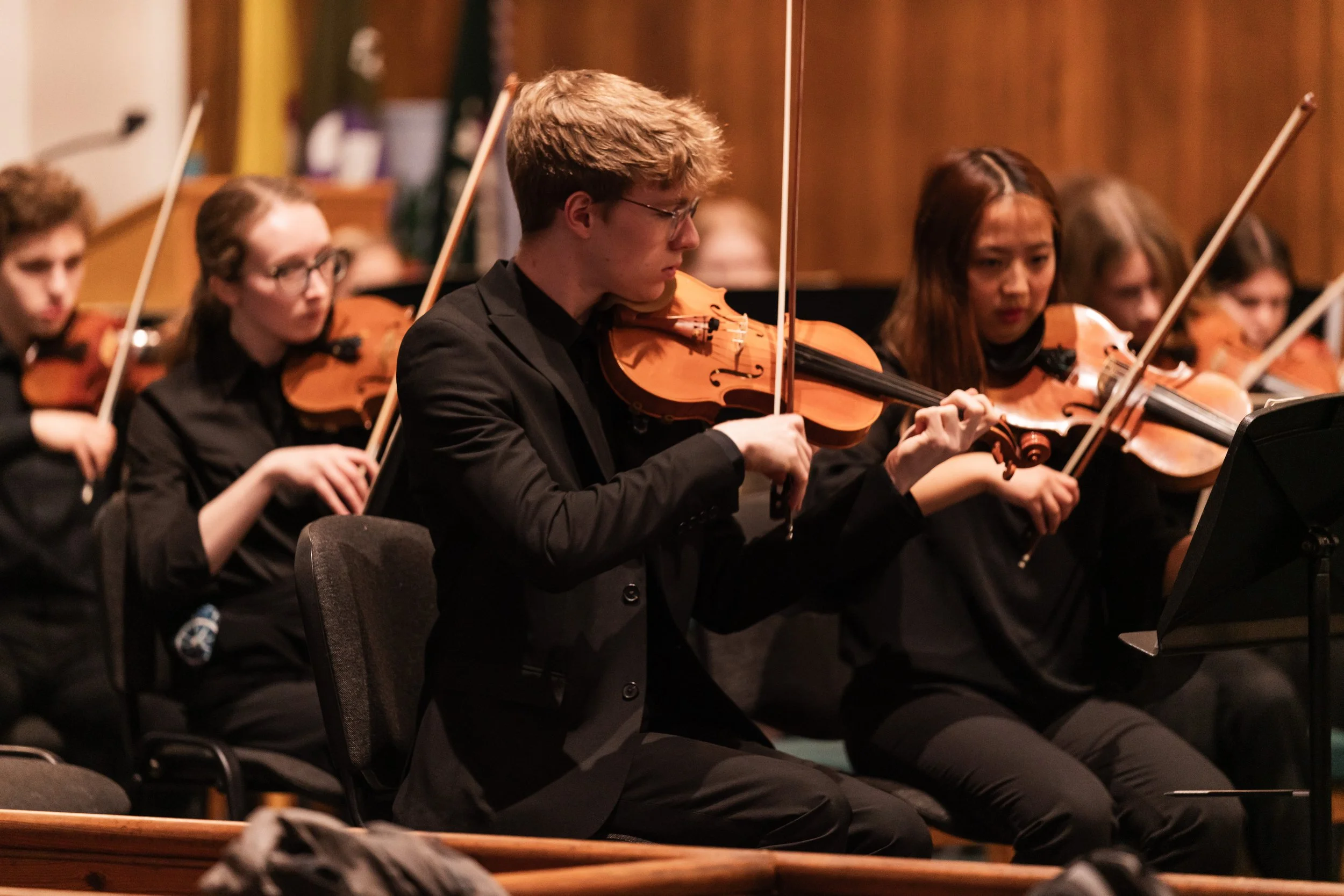 Young musicians in black attire playing violins during an orchestral performance in a concert hall.