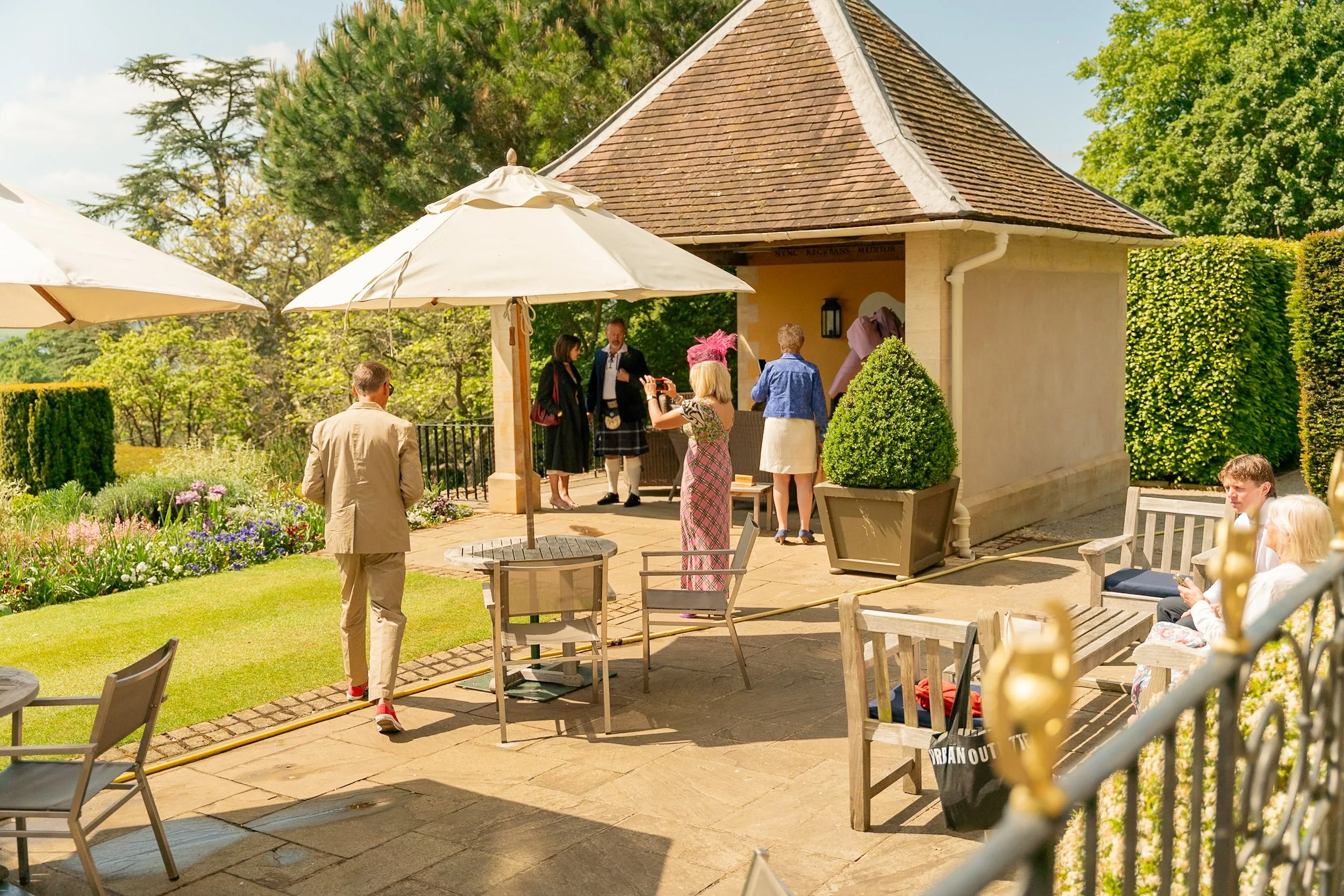 People gathered outdoors in a garden patio for a celebration, with some taking photos and others sitting on benches under umbrellas, near a small building with a brown roof, greenery, and flower beds.