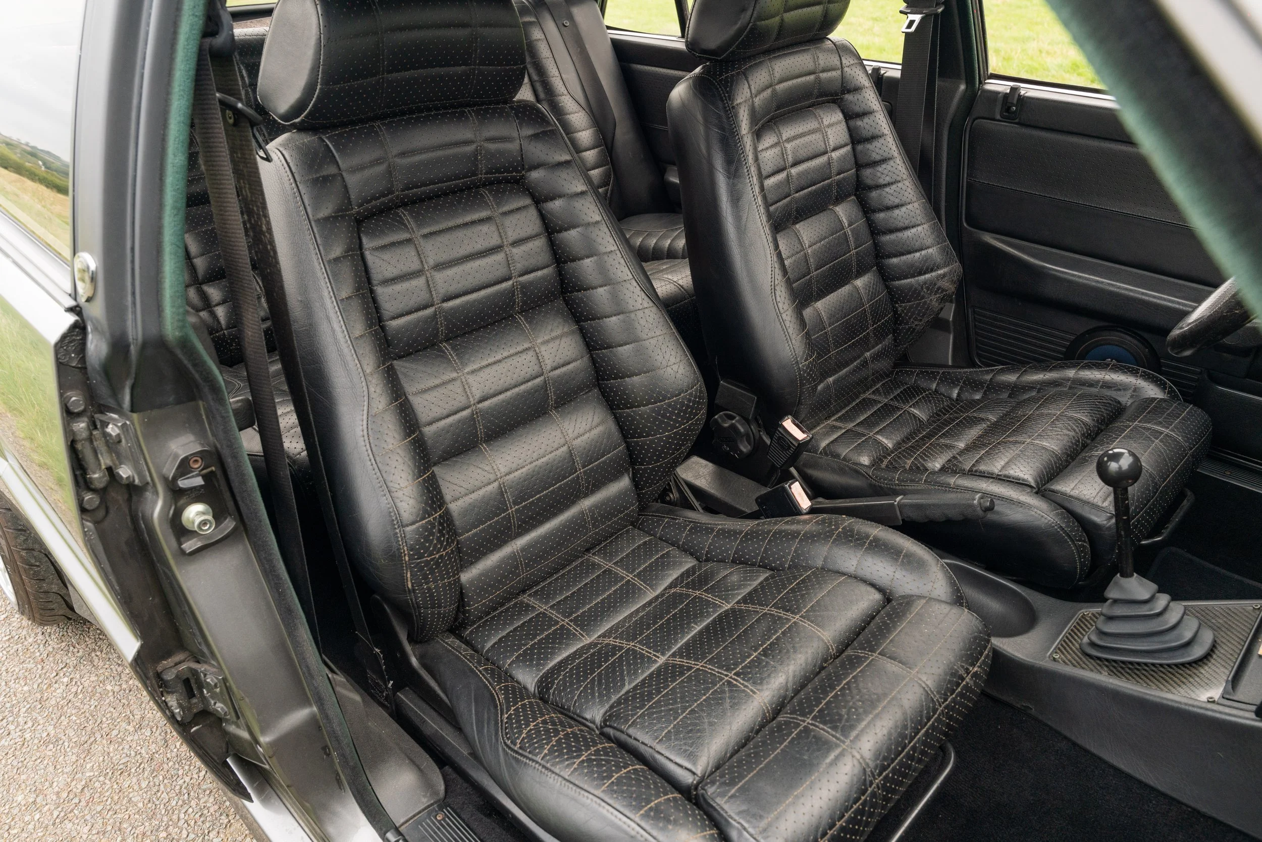 The interior of a classic car showing black leather seats with yellow stitching, manual gear shift, and dashboard.